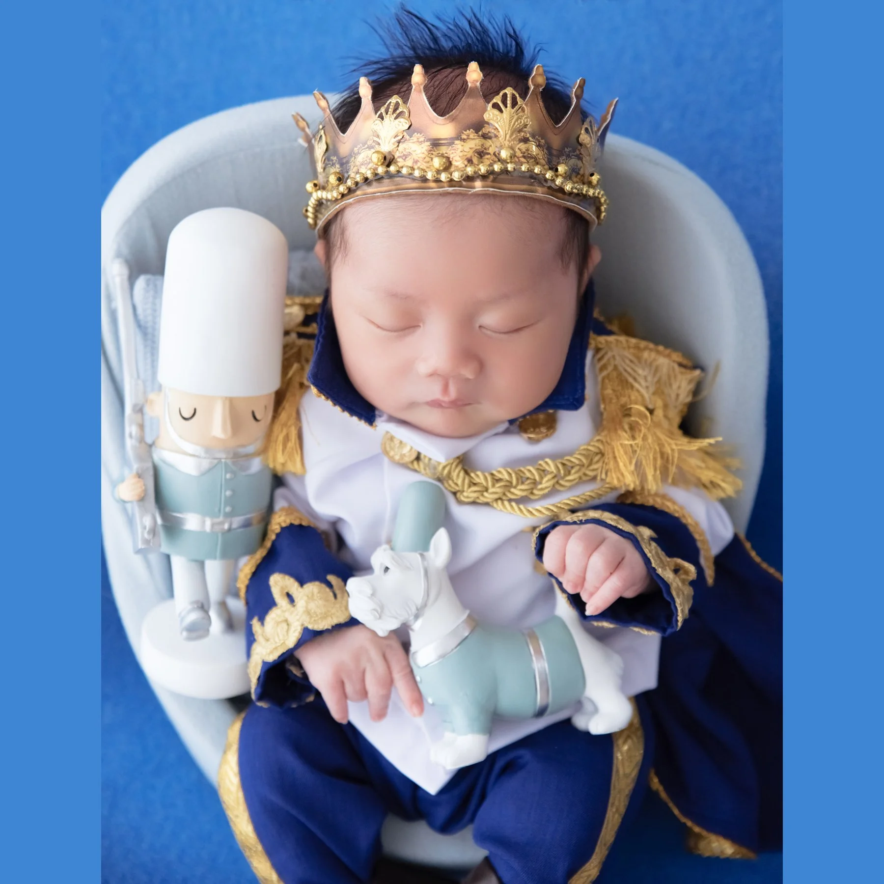 Close-up of a sleeping newborn in a gold crown and prince uniform with a toy soldier resting on the lap