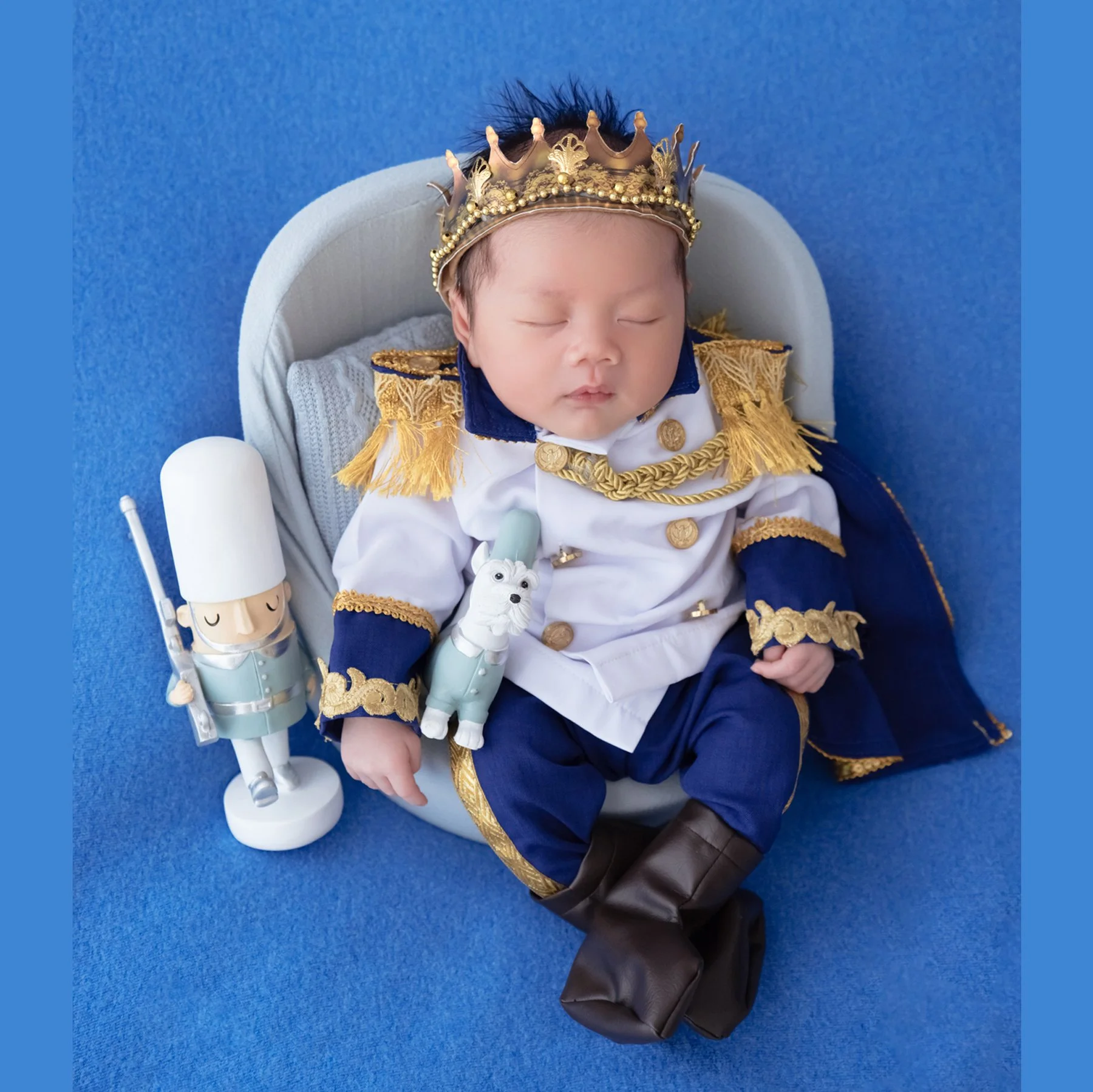 Sleeping newborn wearing a gold crown and blue-and-gold prince uniform in a small seat beside toy soldiers