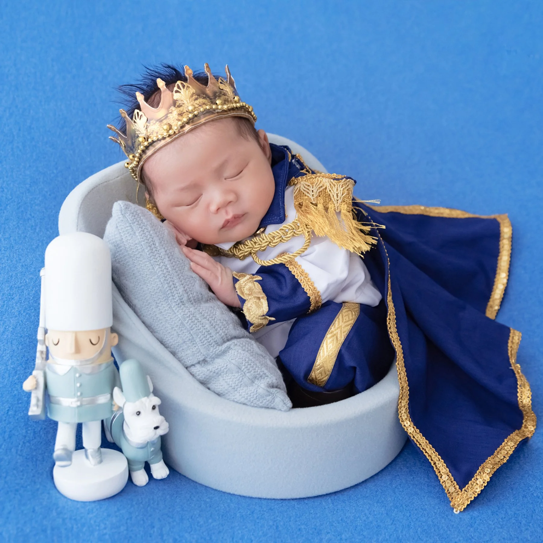 Sleeping newborn wearing a gold crown and blue royal cape, curled in a grey basket beside toy soldiers
