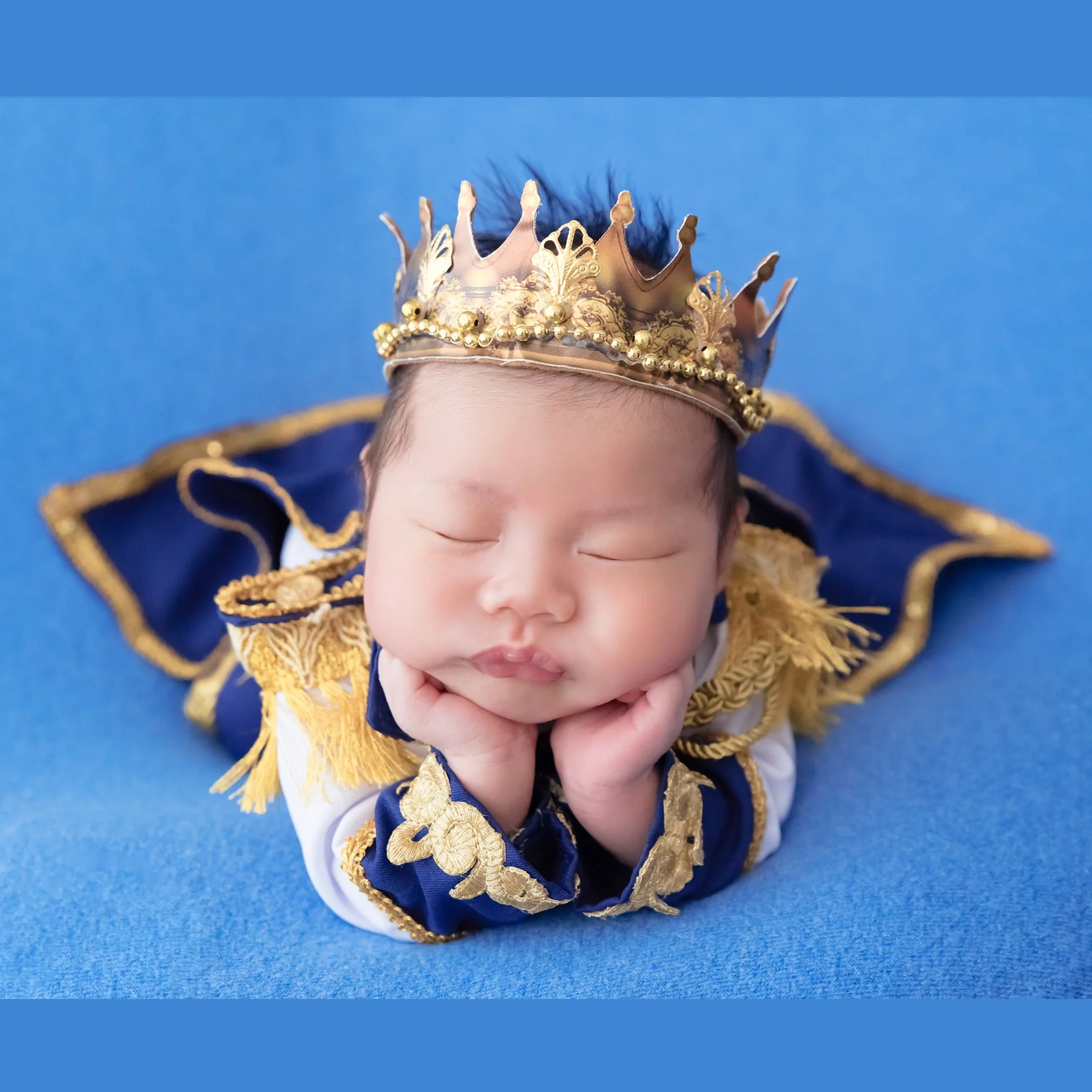 Sleeping newborn in a gold crown and blue prince cape resting the chin on folded hands against a blue backdrop
