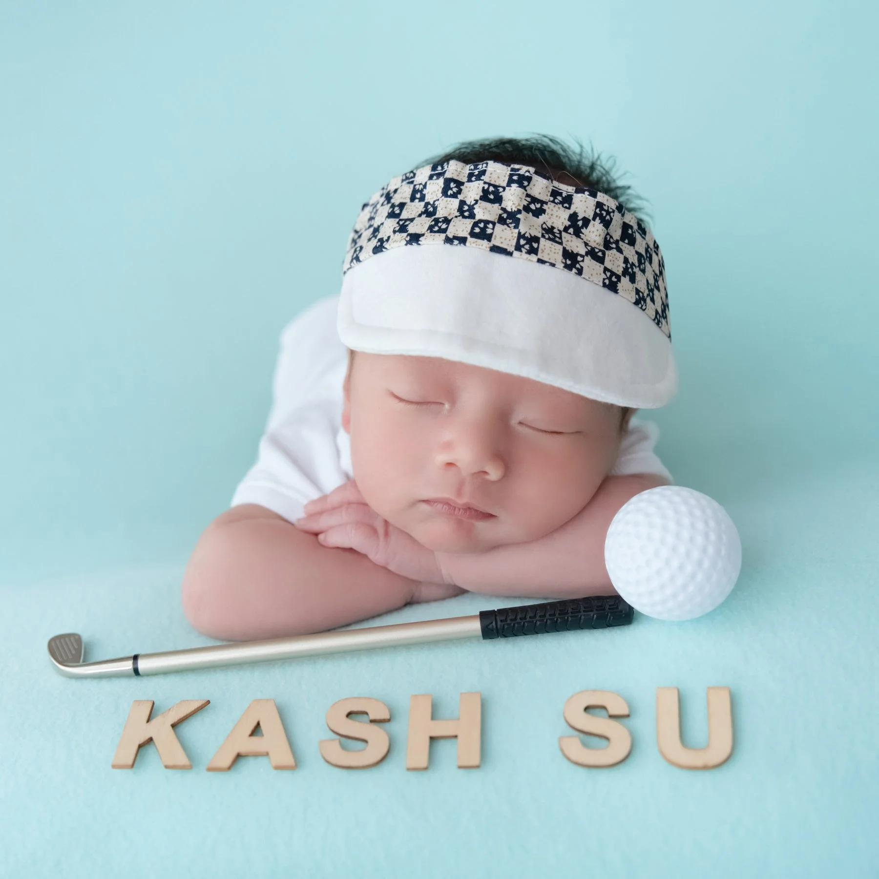 Close-up of a sleeping newborn in a white golf shirt and checked visor resting the chin on folded arms beside a miniature golf club and ball