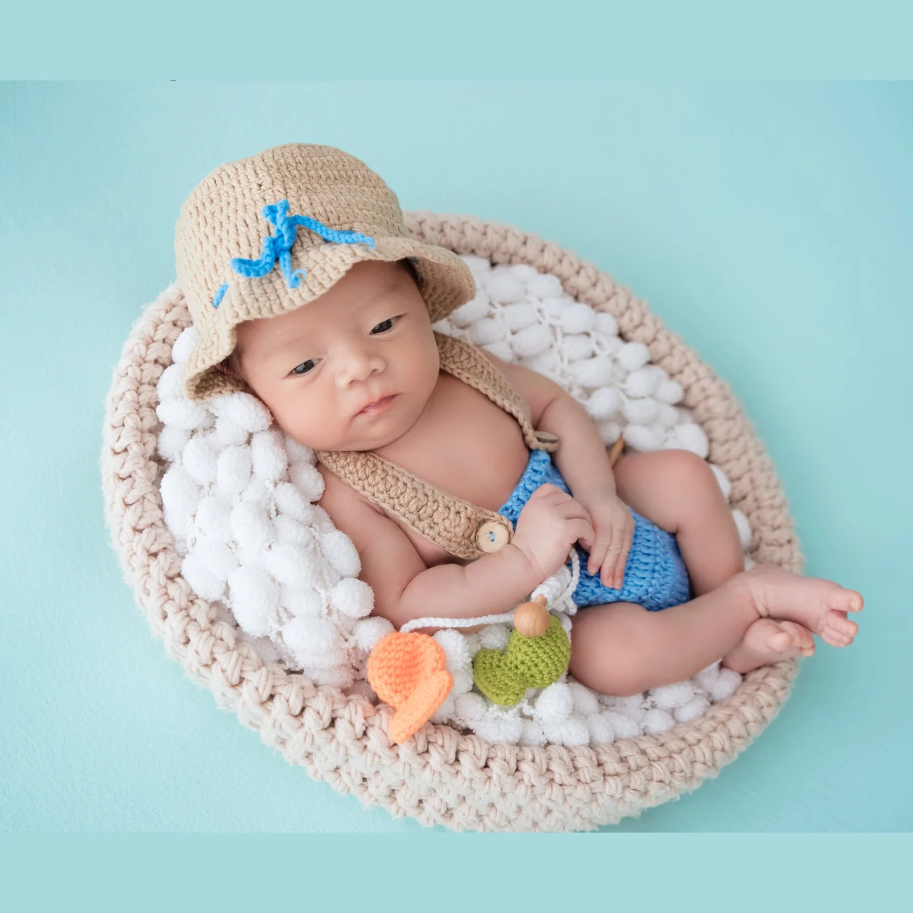 Awake newborn in a straw hat and blue knitted shorts reclining in a white-filled basket with a toy fishing rod and colourful fish