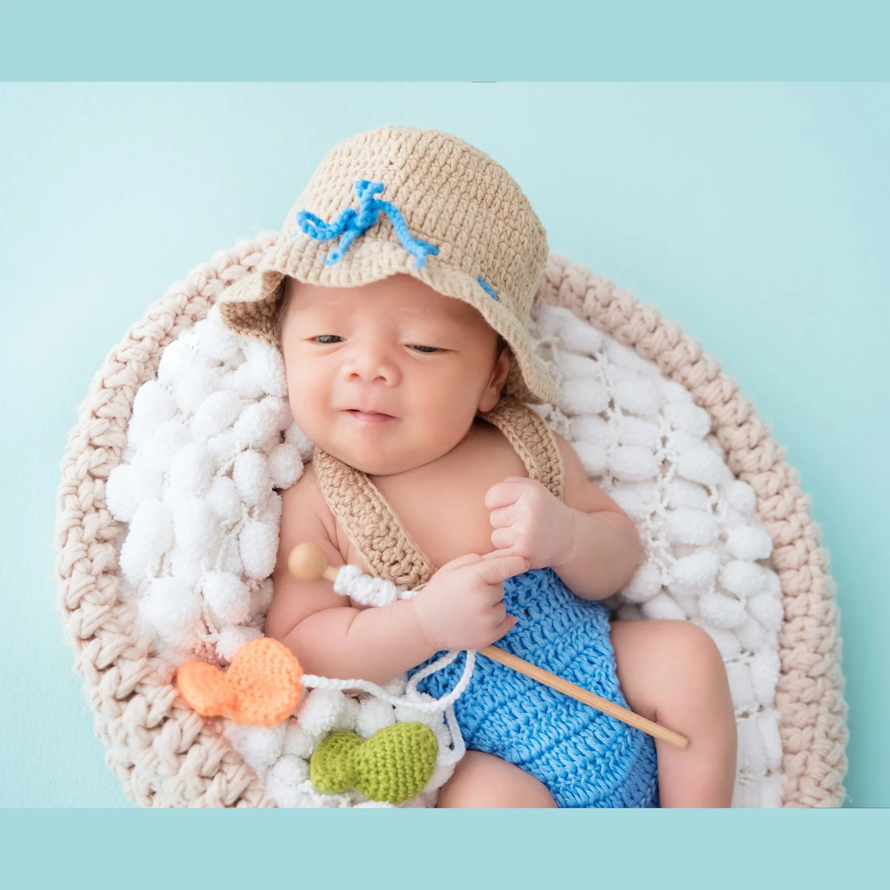 Close-up of an awake newborn in a straw hat and blue knitted shorts holding a toy fishing rod in a white-filled basket