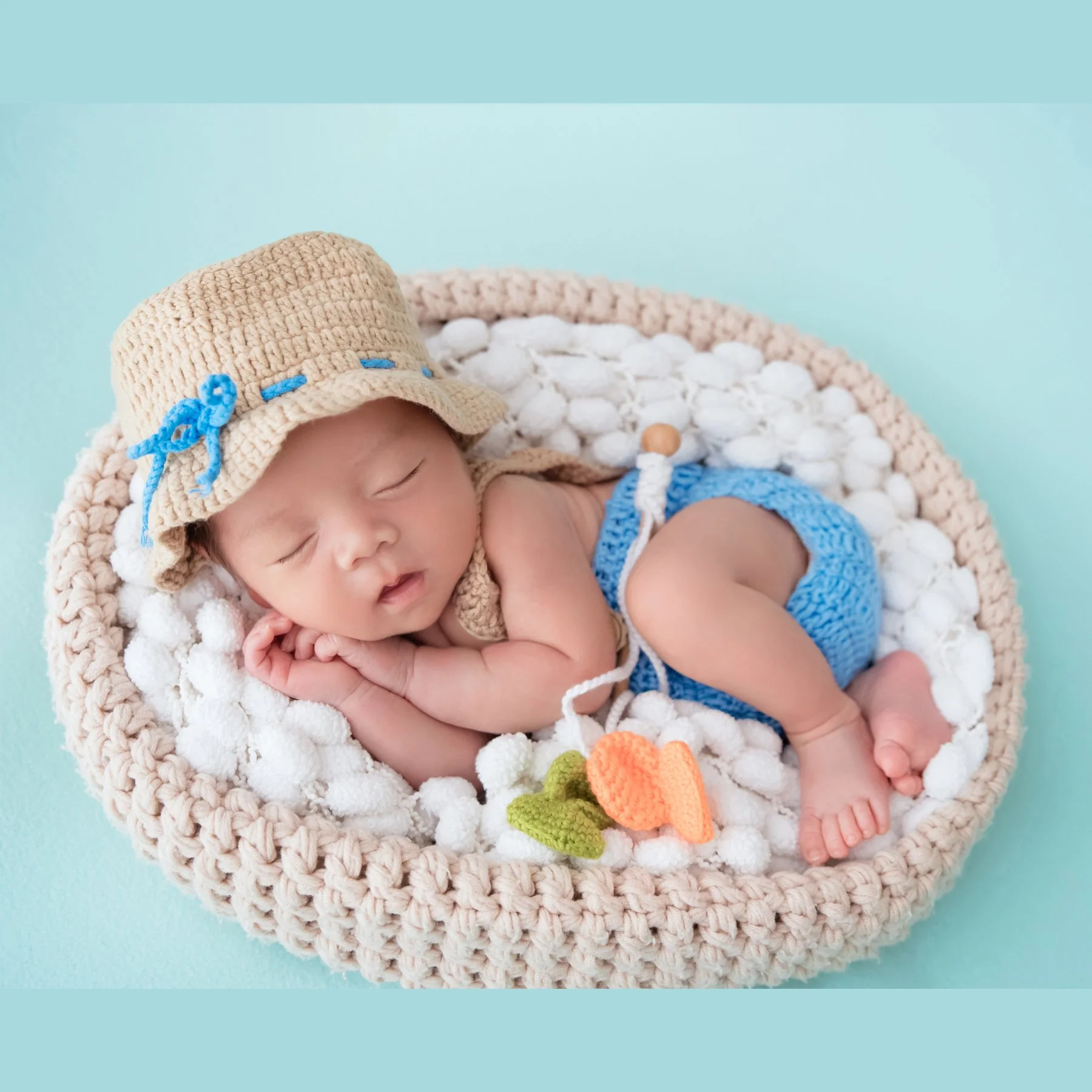 Sleeping newborn in a straw hat and blue knitted shorts curled on one side in a white-filled basket beside a toy fishing rod and fish