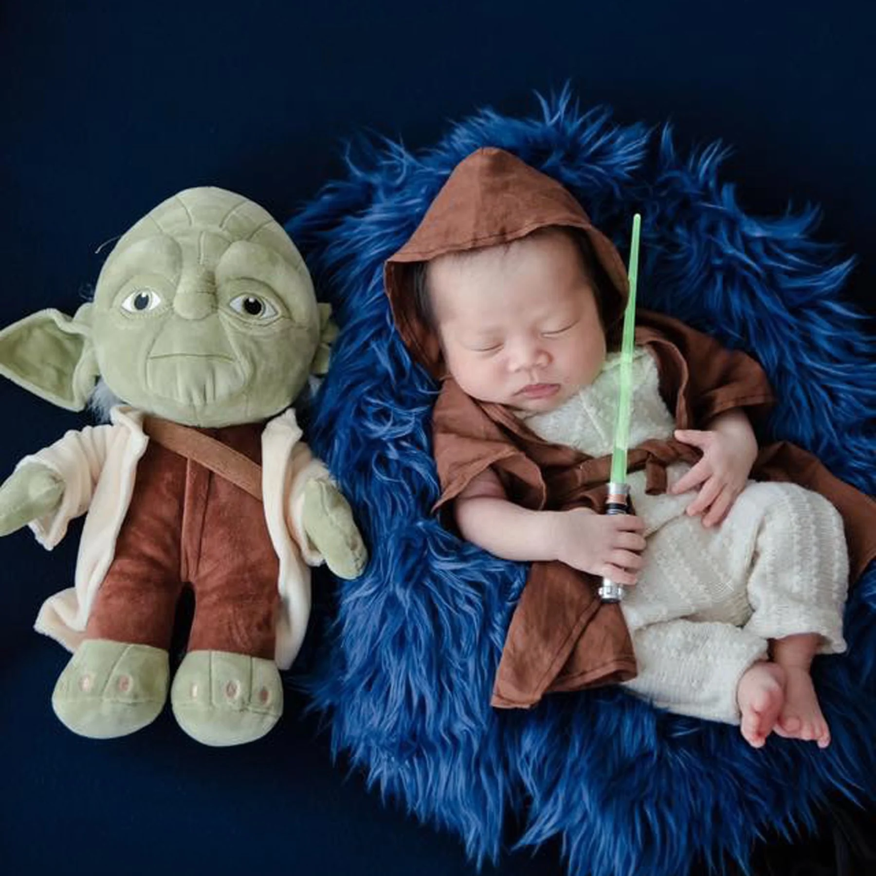 Sleeping newborn in a brown Jedi robe holding a green toy lightsaber beside a Yoda plush on blue fur