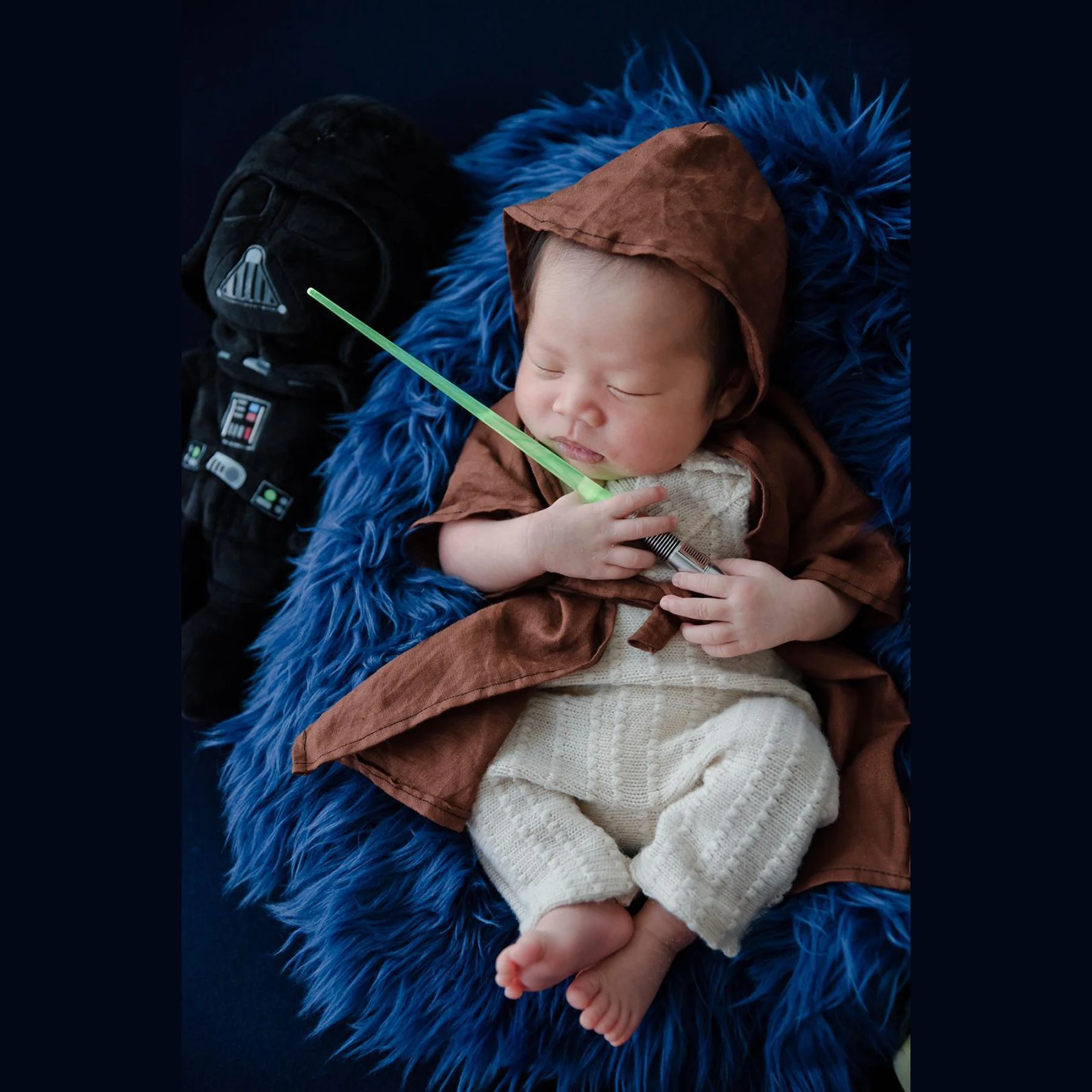 Sleeping newborn in a hooded Jedi robe holding a green toy lightsaber beside a Darth Vader plush