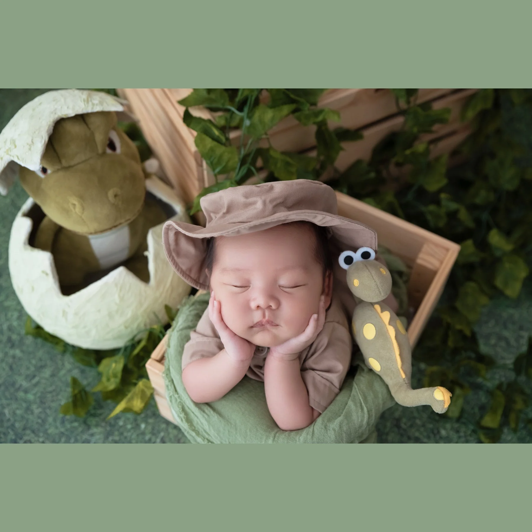 Sleeping newborn in a safari hat and green wrap resting both cheeks on hands in a wooden crate beside dinosaur plush toys