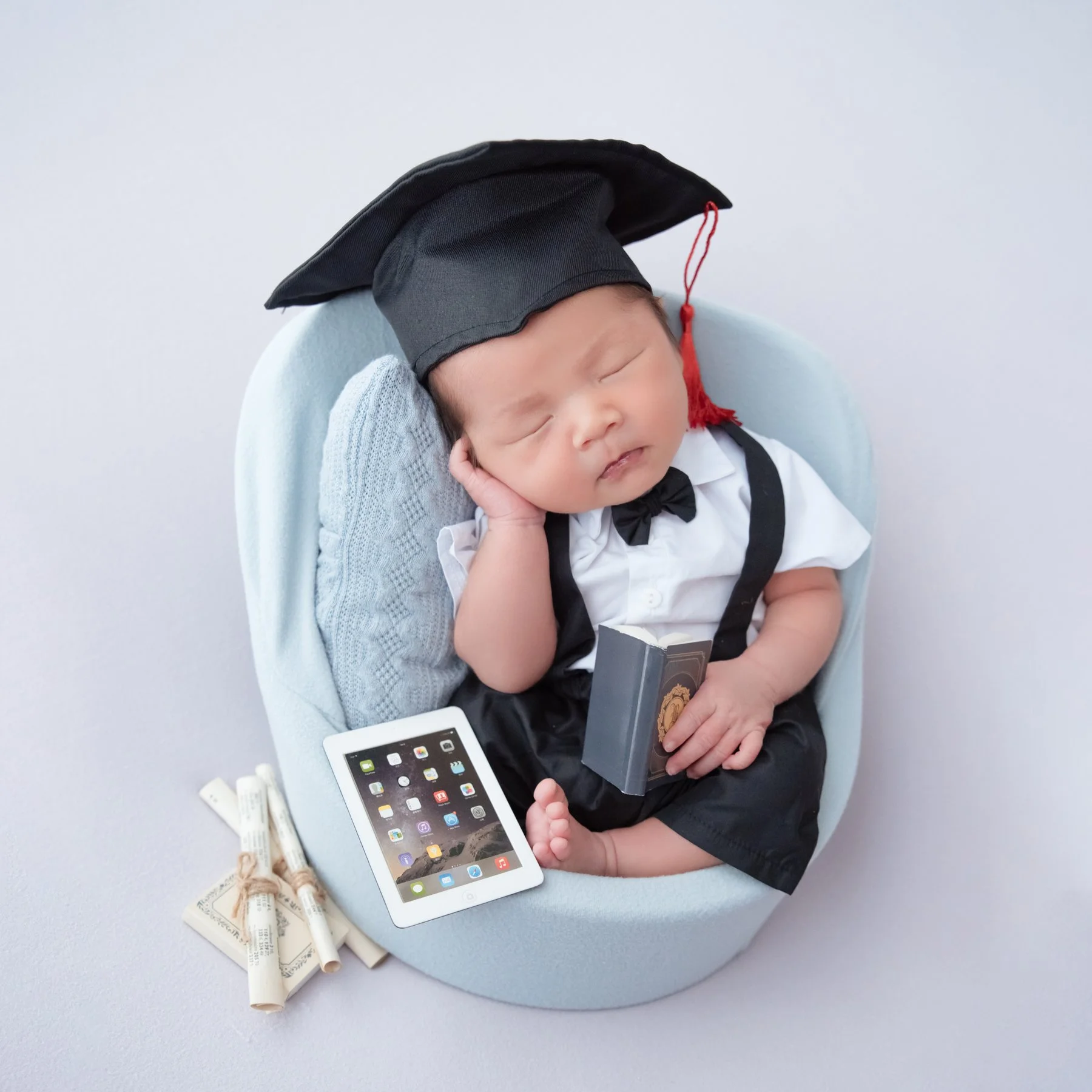 Sleeping newborn in a graduation cap, white shirt and black bow tie seated in a pale blue basket beside rolled certificates