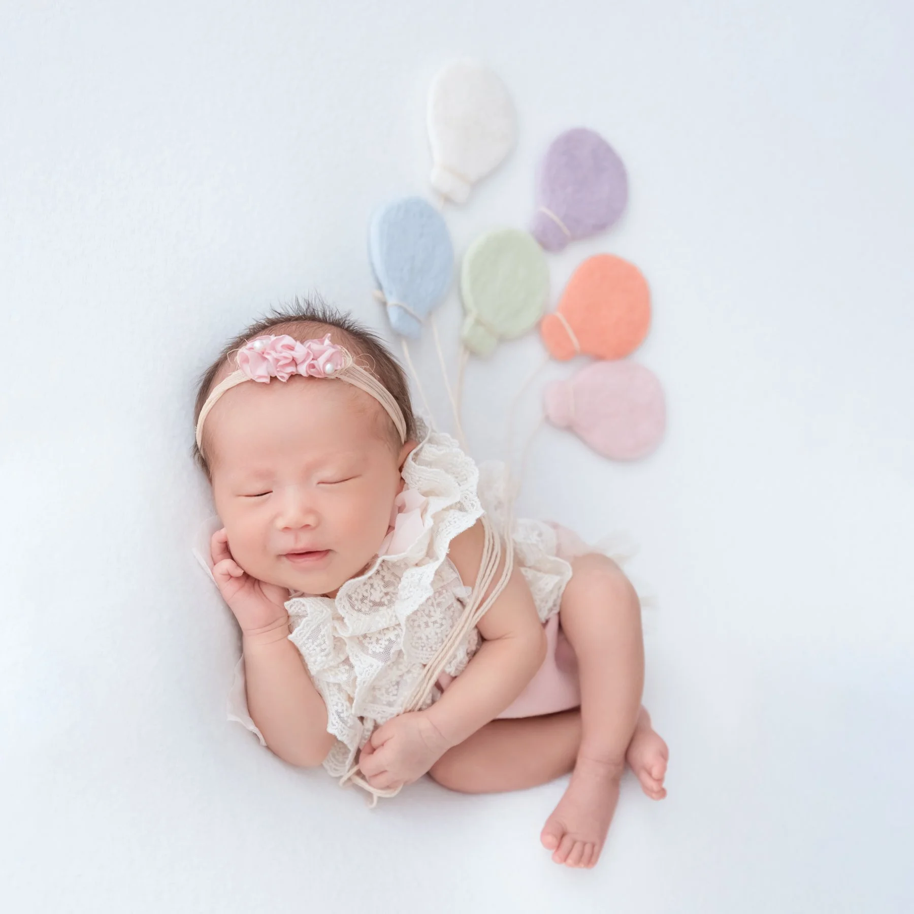 Smiling sleeping newborn in a white lace romper and pink floral headband curled on one side beside pastel balloon decorations