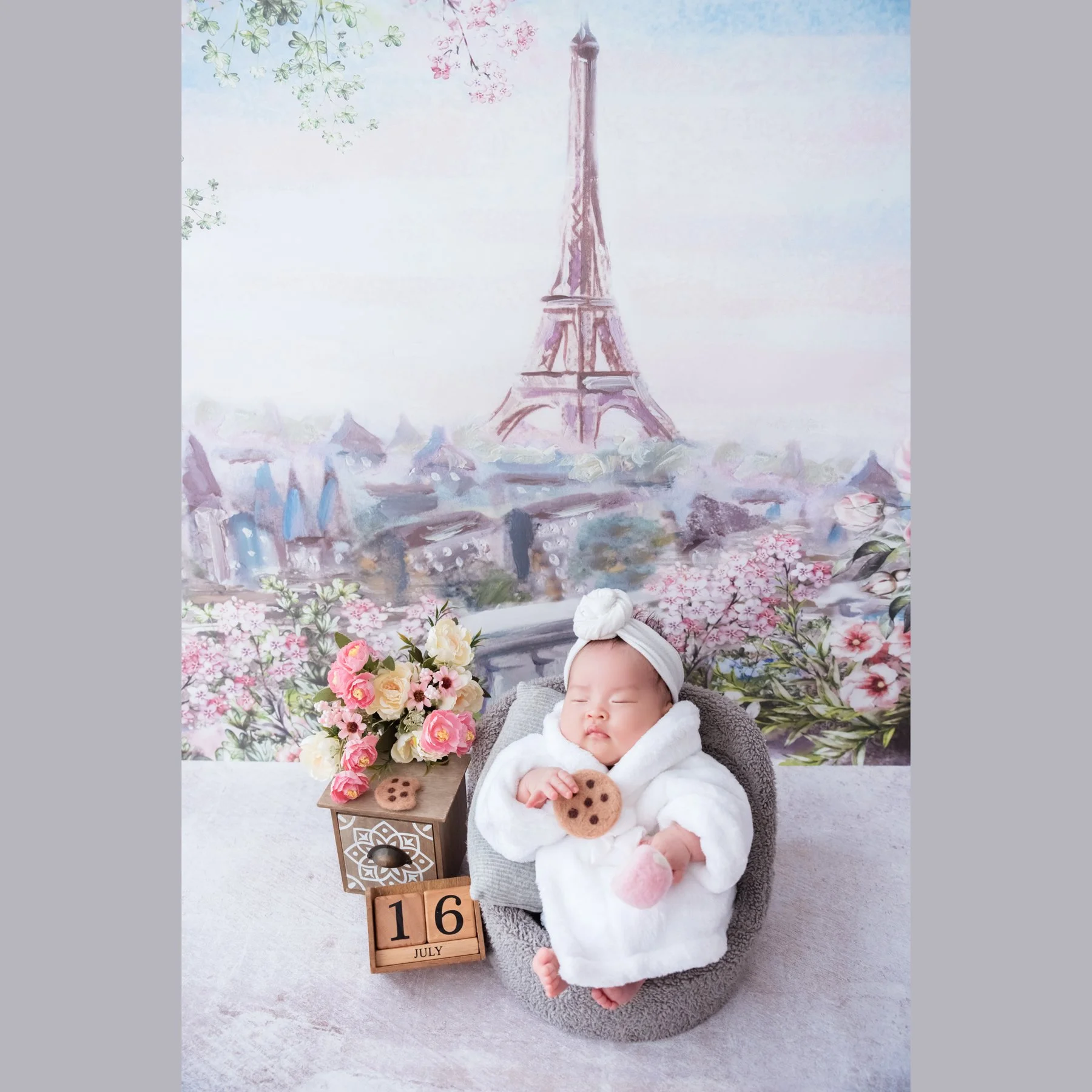 Sleeping newborn in a white dress and bow headband reclining in a grey basket with a toy biscuit beside flowers against a Paris Eiffel Tower backdrop