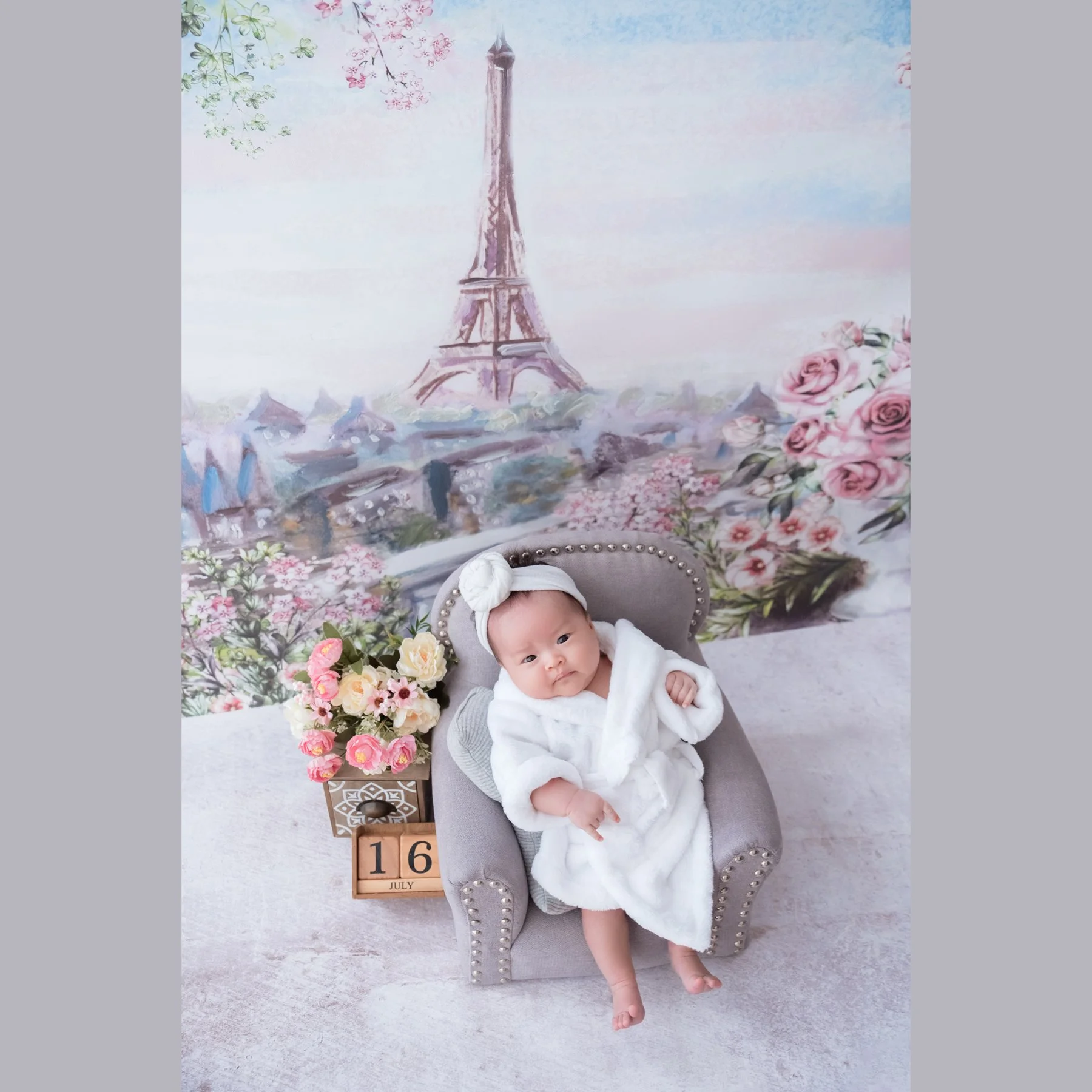 Awake newborn in a white dress and bow headband reclining in a miniature grey armchair beside flowers against a Paris Eiffel Tower backdrop