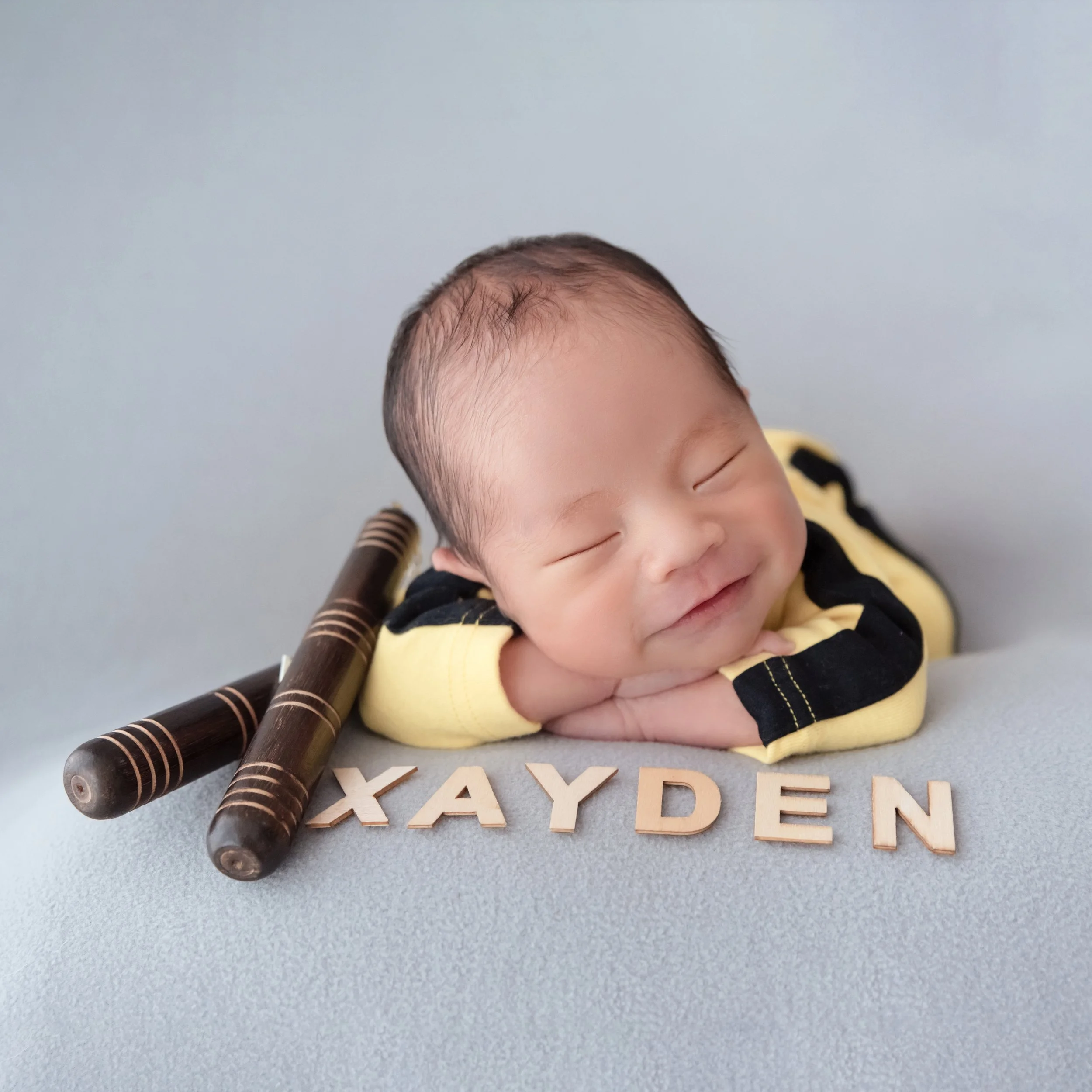 Smiling sleeping newborn in a yellow-and-black martial arts outfit resting the chin on folded arms beside toy nunchucks