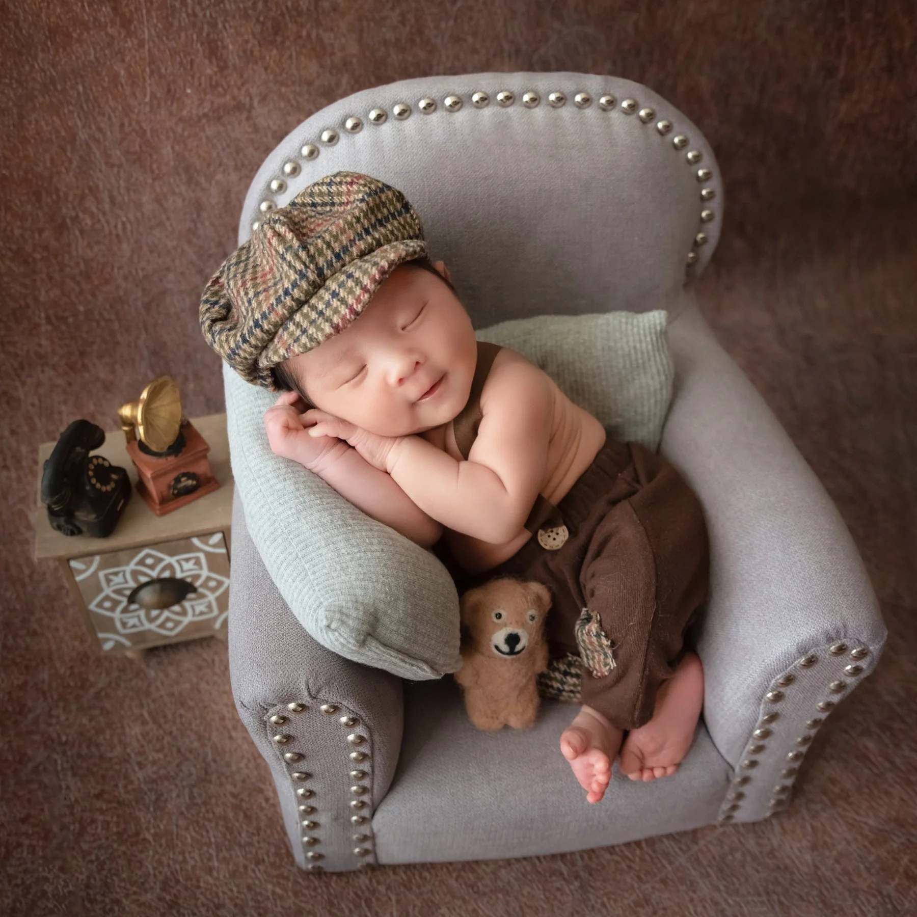 Sleeping newborn in a checked cap and brown wrap resting the cheek on folded hands in a miniature grey armchair beside a teddy bear and vintage telephone