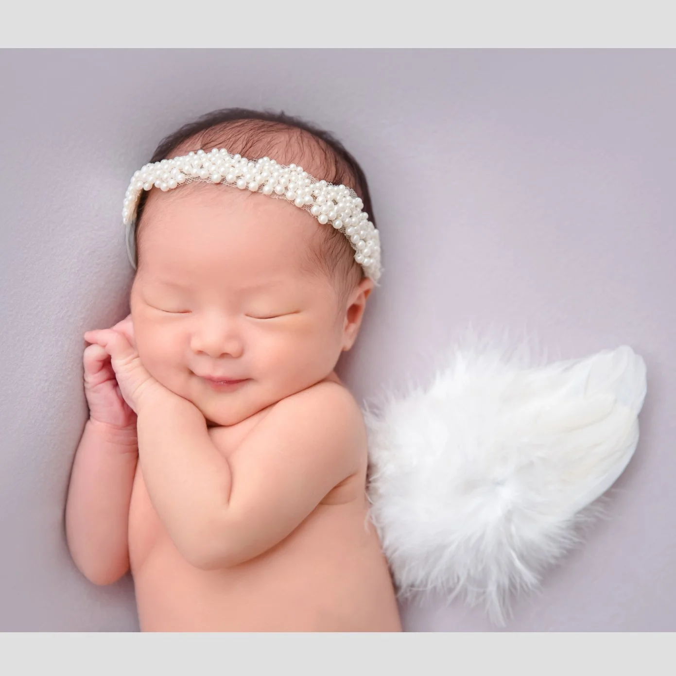 Smiling newborn wearing a white lace headband with hands tucked beside the cheek and white angel wings behind
