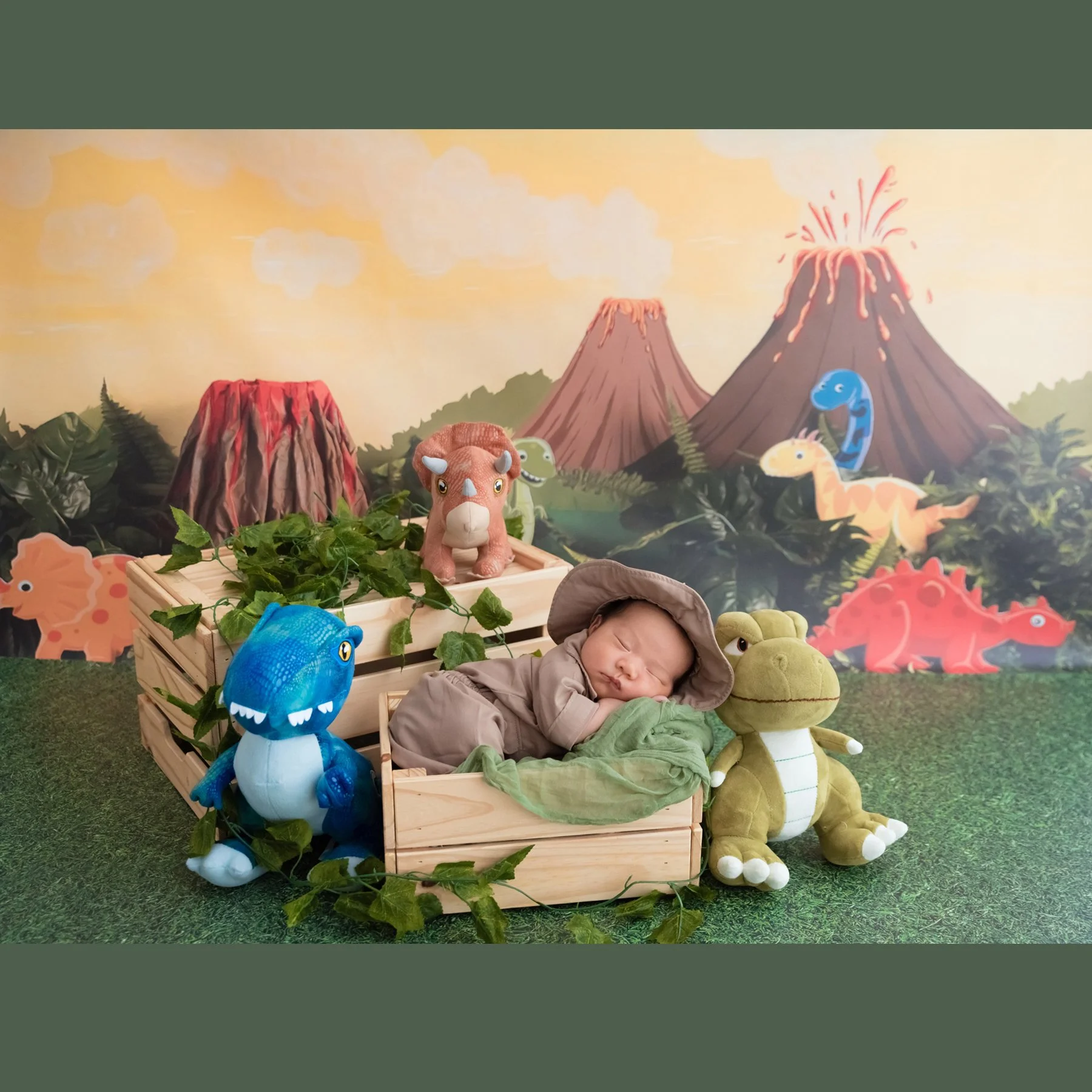 Sleeping newborn in a safari hat and green wrap curled in a wooden crate among dinosaur plush toys against a volcano backdrop