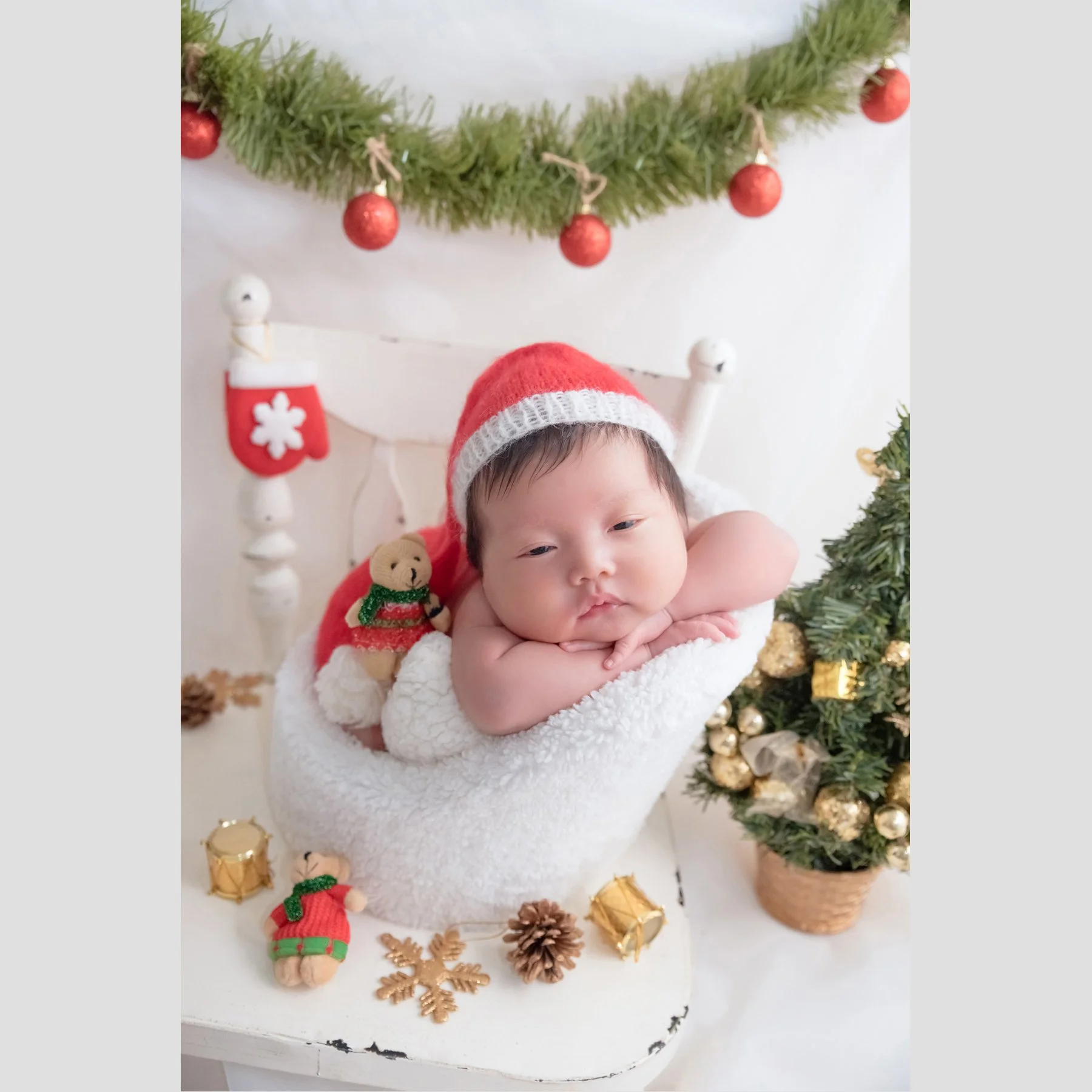 Awake newborn in a red Santa hat resting the chin on folded arms in a white basket beneath a Christmas garland and red baubles