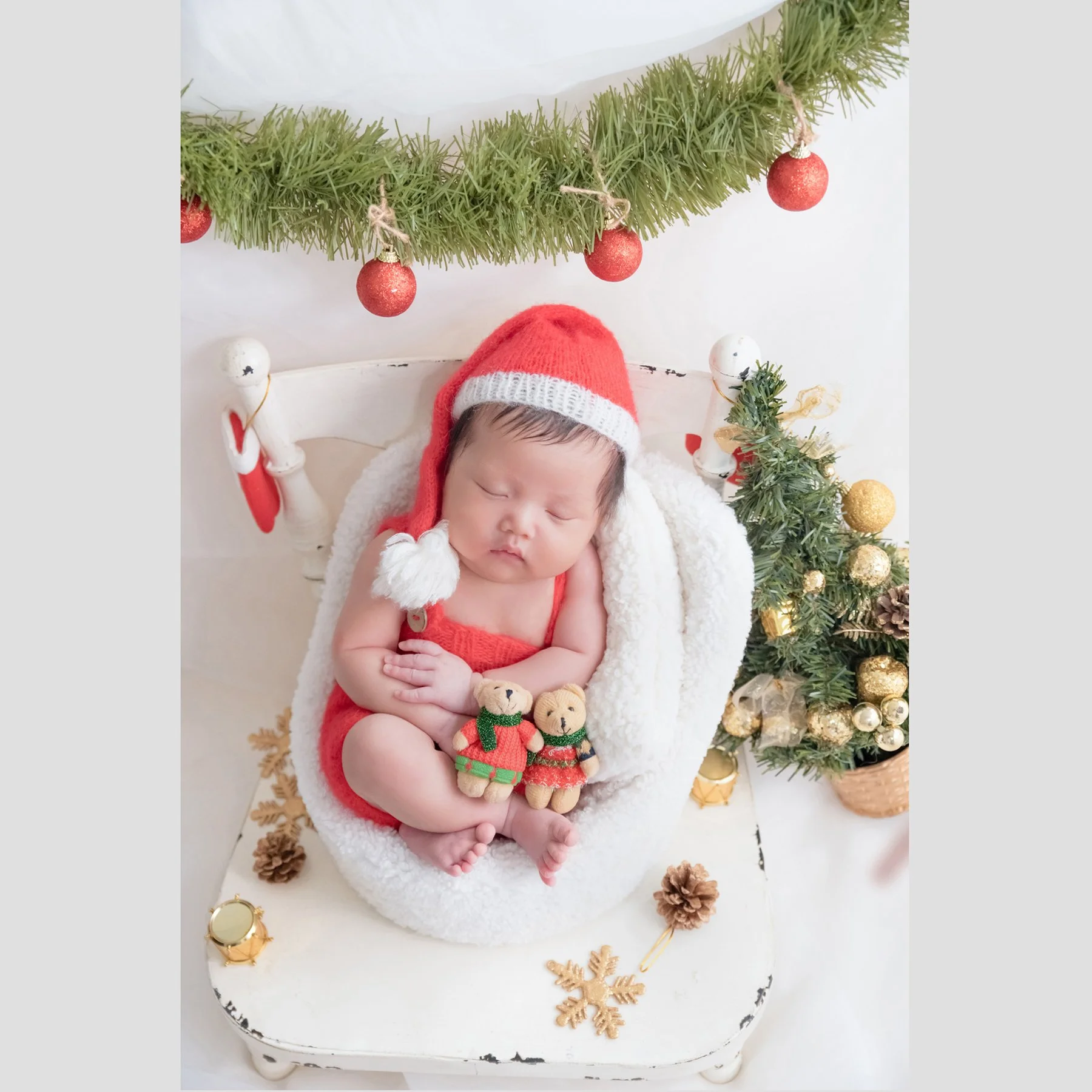 Sleeping newborn in a red Santa hat and outfit curled in a white basket with a teddy bear beside a miniature Christmas tree and gold decorations