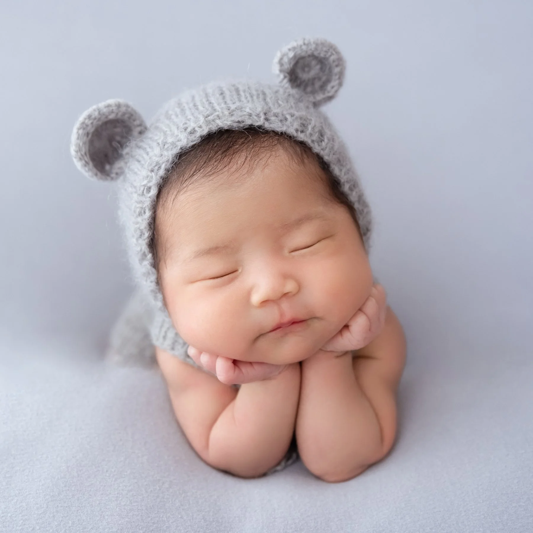 Sleeping newborn wearing a grey bear hat resting the chin on both hands against a pale blue backdrop