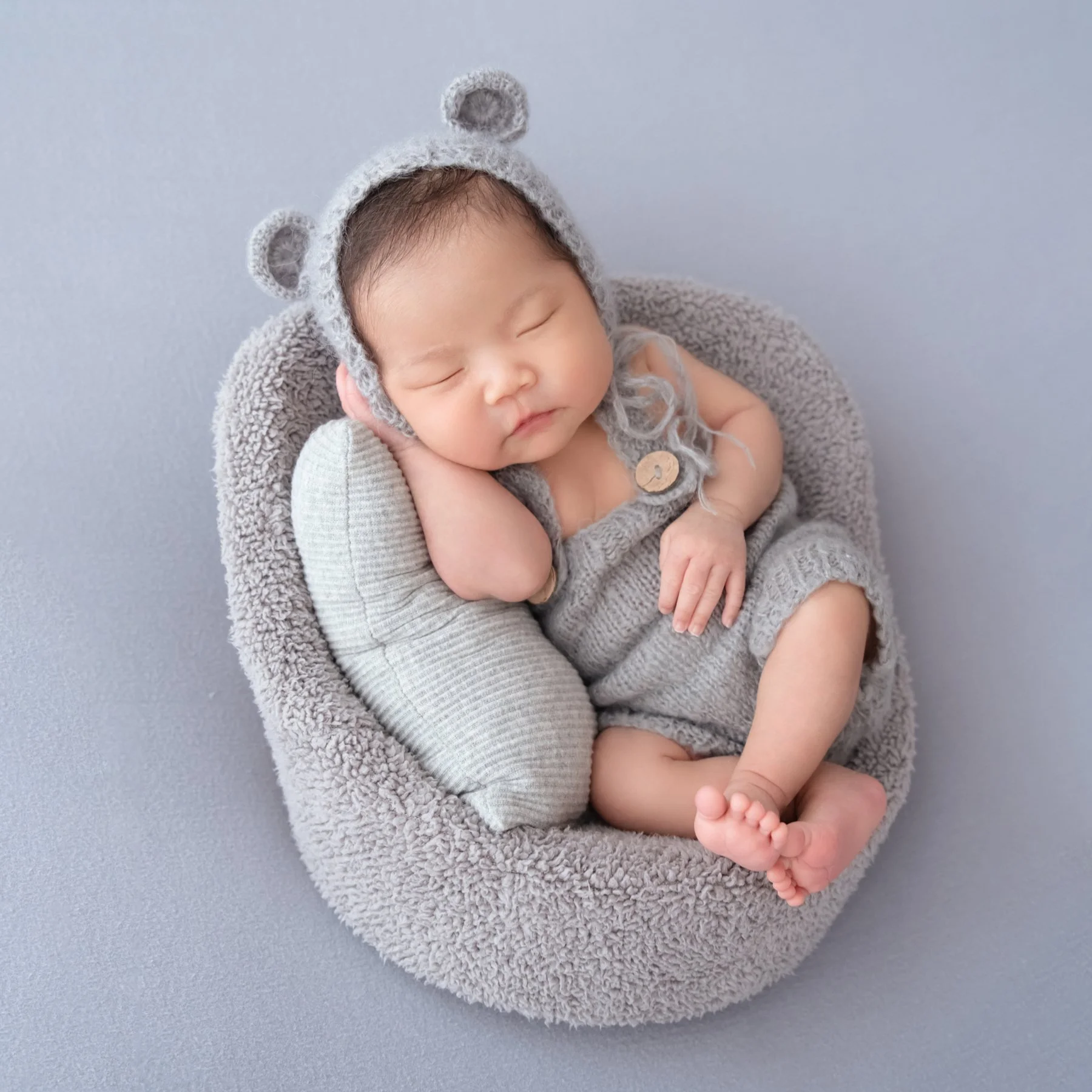 Sleeping newborn in a grey knitted bear hat and romper curled in a grey basket with a pale cushion