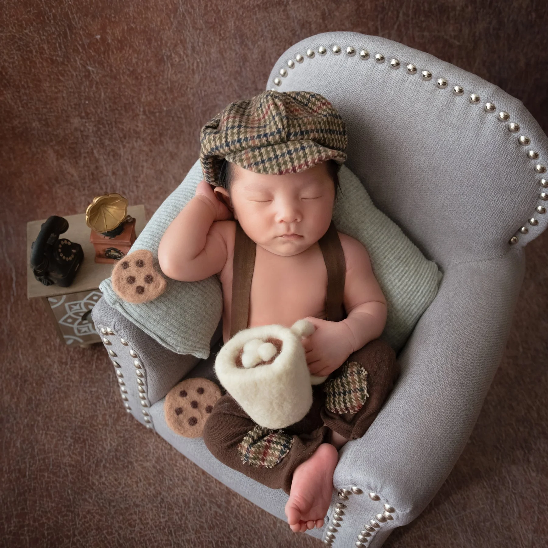 Sleeping newborn in a checked cap and brown wrap reclining with hands beside the head in a miniature grey armchair, with toy cookies and a white cup