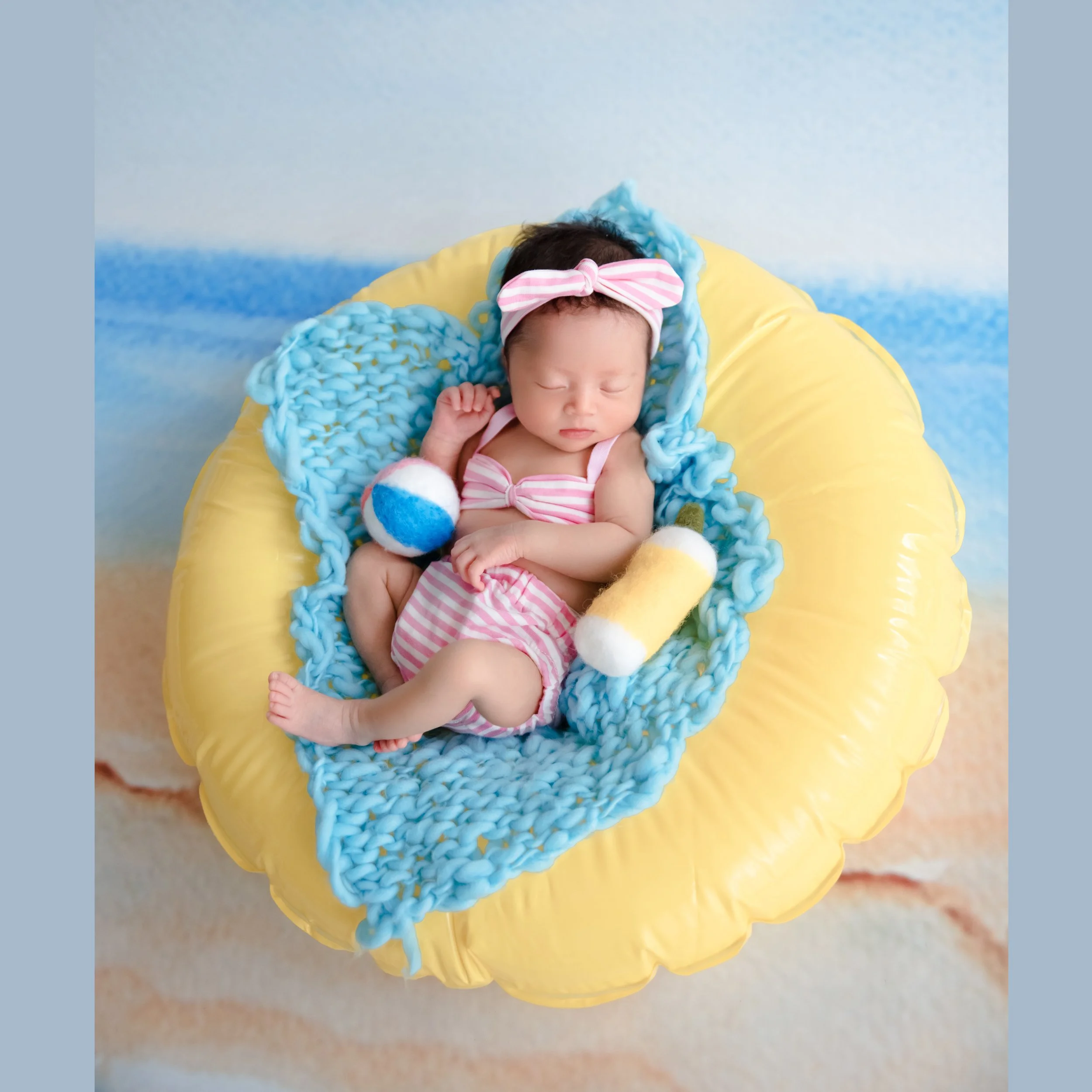 Sleeping newborn in a striped pink swimsuit and bow headband reclining on a blue blanket in a yellow inflatable ring beside a toy beach ball