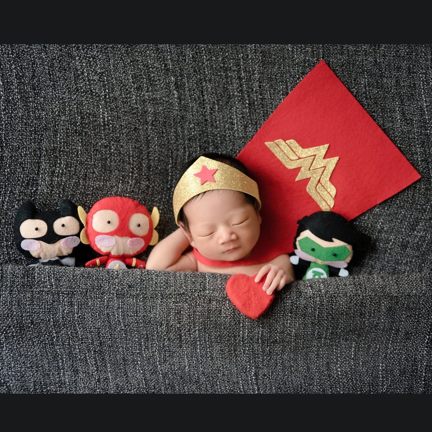 Sleeping newborn wearing a Wonder Woman headband beside a red superhero cushion and plush toys on a grey backdrop