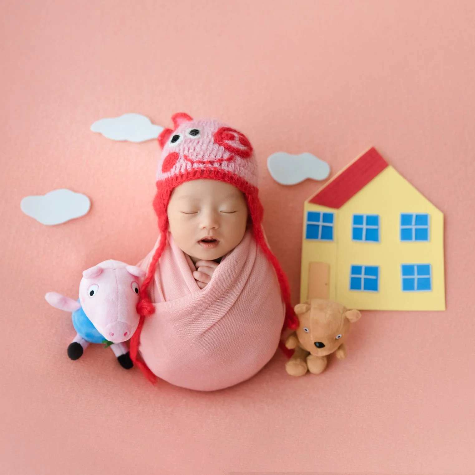 Pink-swaddled newborn wearing a Peppa Pig hat beside a toy pig and a yellow house on a pink backdrop