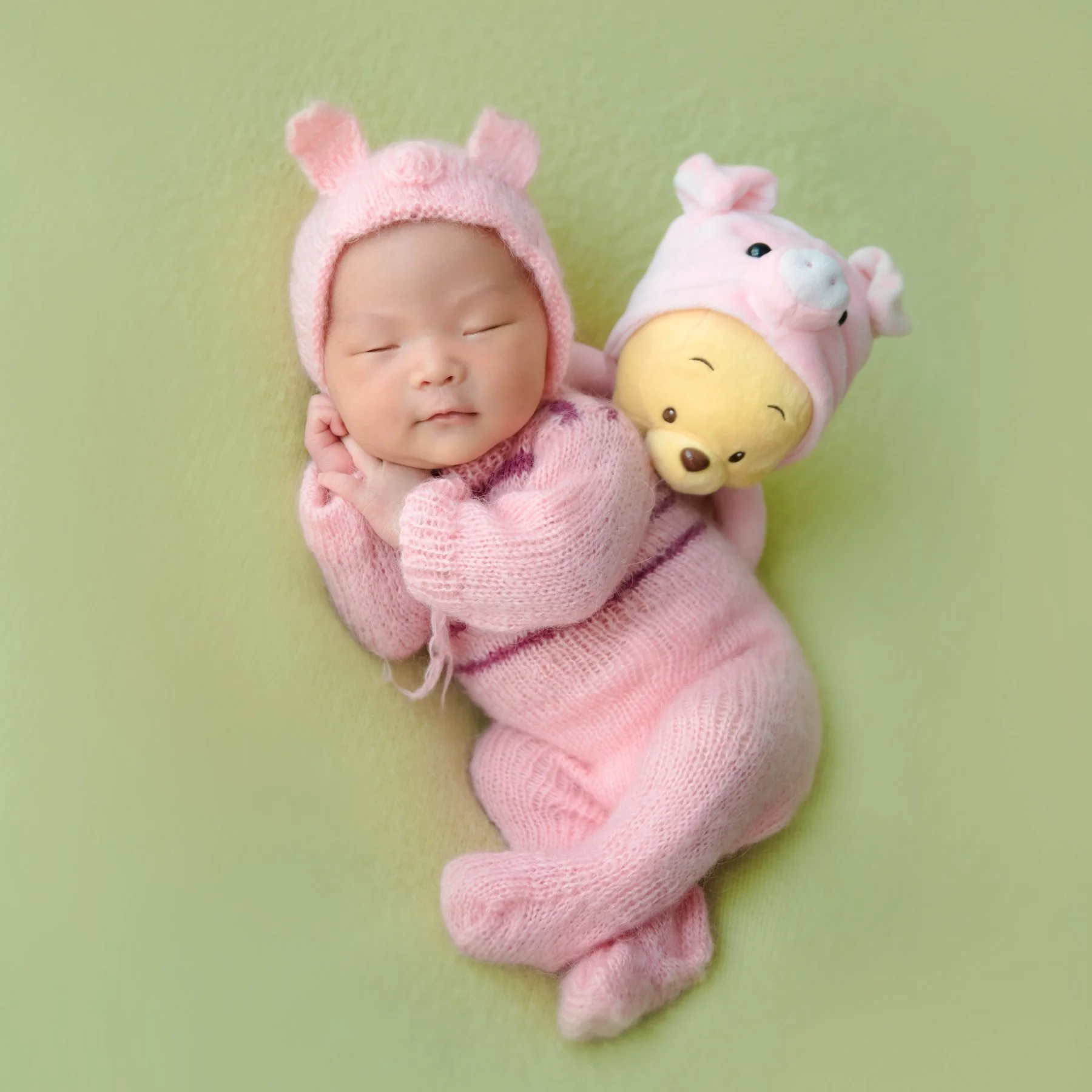Sleeping newborn in a pink knitted Piglet outfit lying on one side beside a Winnie the Pooh toy on green