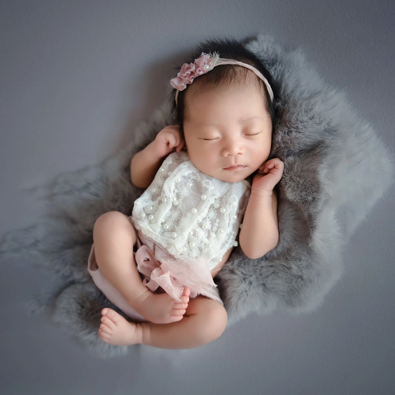 Sleeping newborn in a white lace romper and floral headband reclining with hands beside the head on grey fur