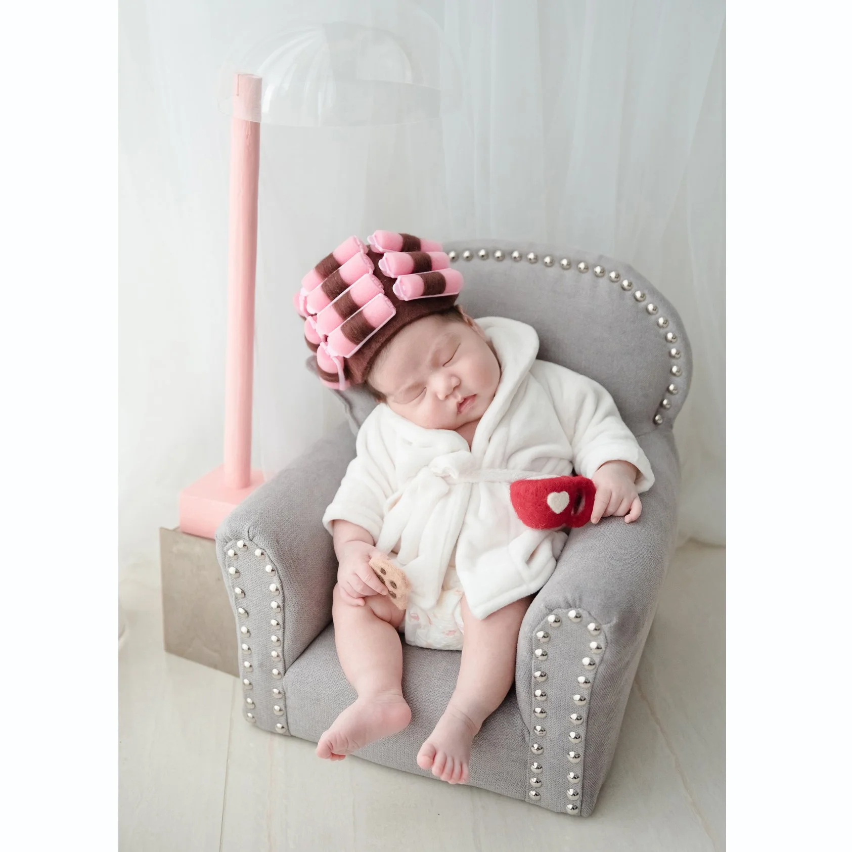 Sleeping newborn in a white bathrobe and pink hair rollers reclining in a miniature grey armchair with a red cup