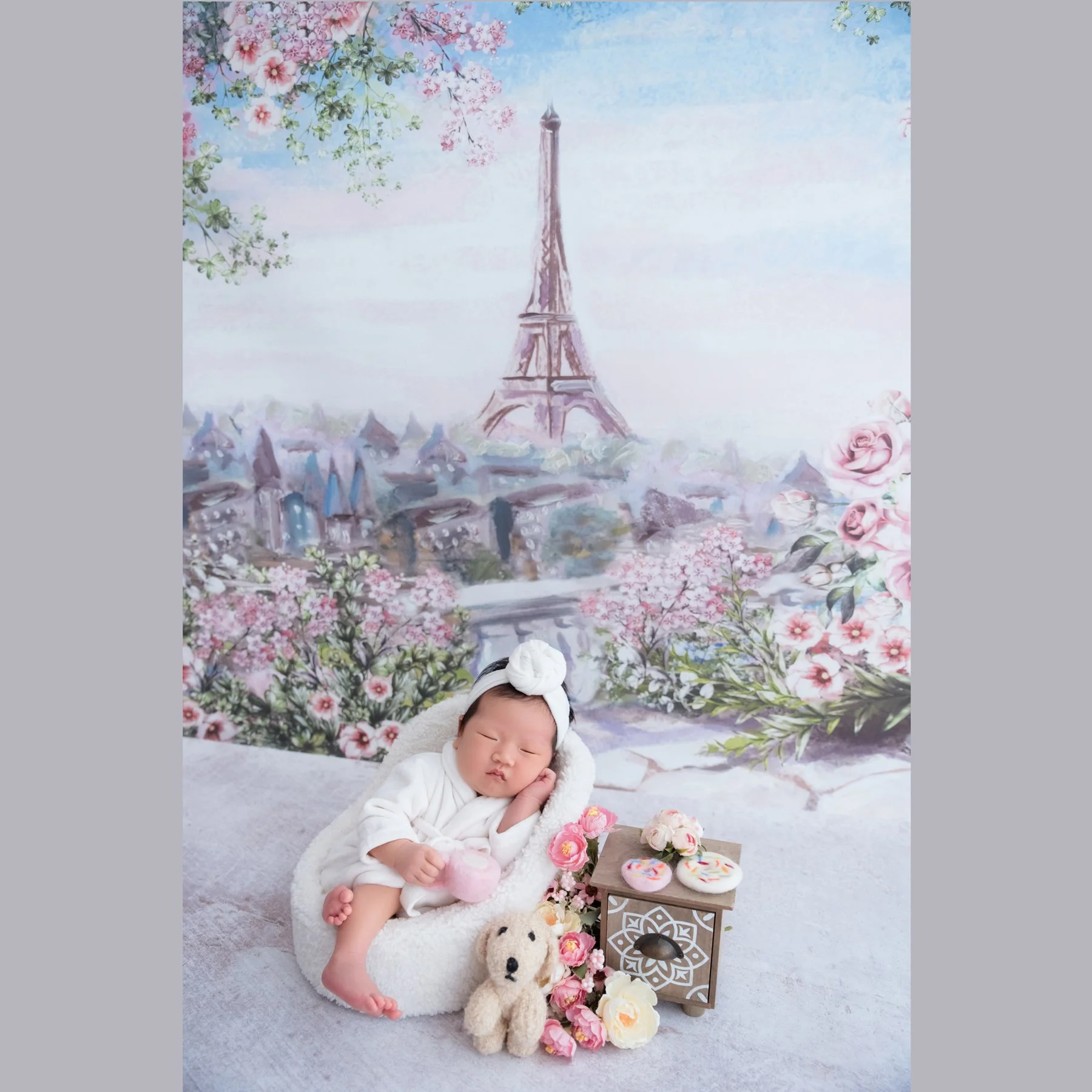 Sleeping newborn in a white dress and bow headband lying on a white blanket beside flowers and a vintage clock against an Eiffel Tower backdrop