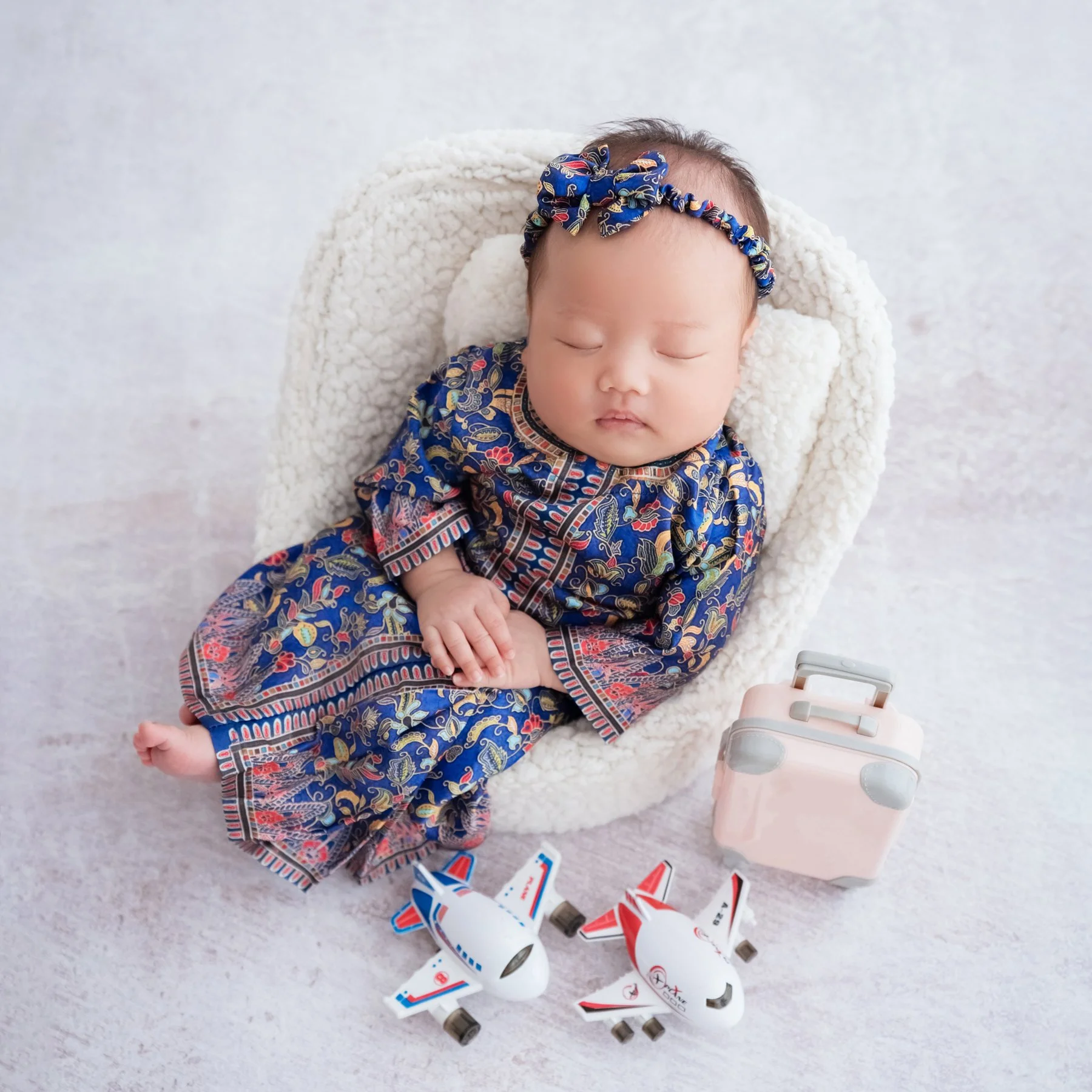 Sleeping newborn in a patterned blue dress and matching headband in a white basket beside toy airplanes and a pink suitcase