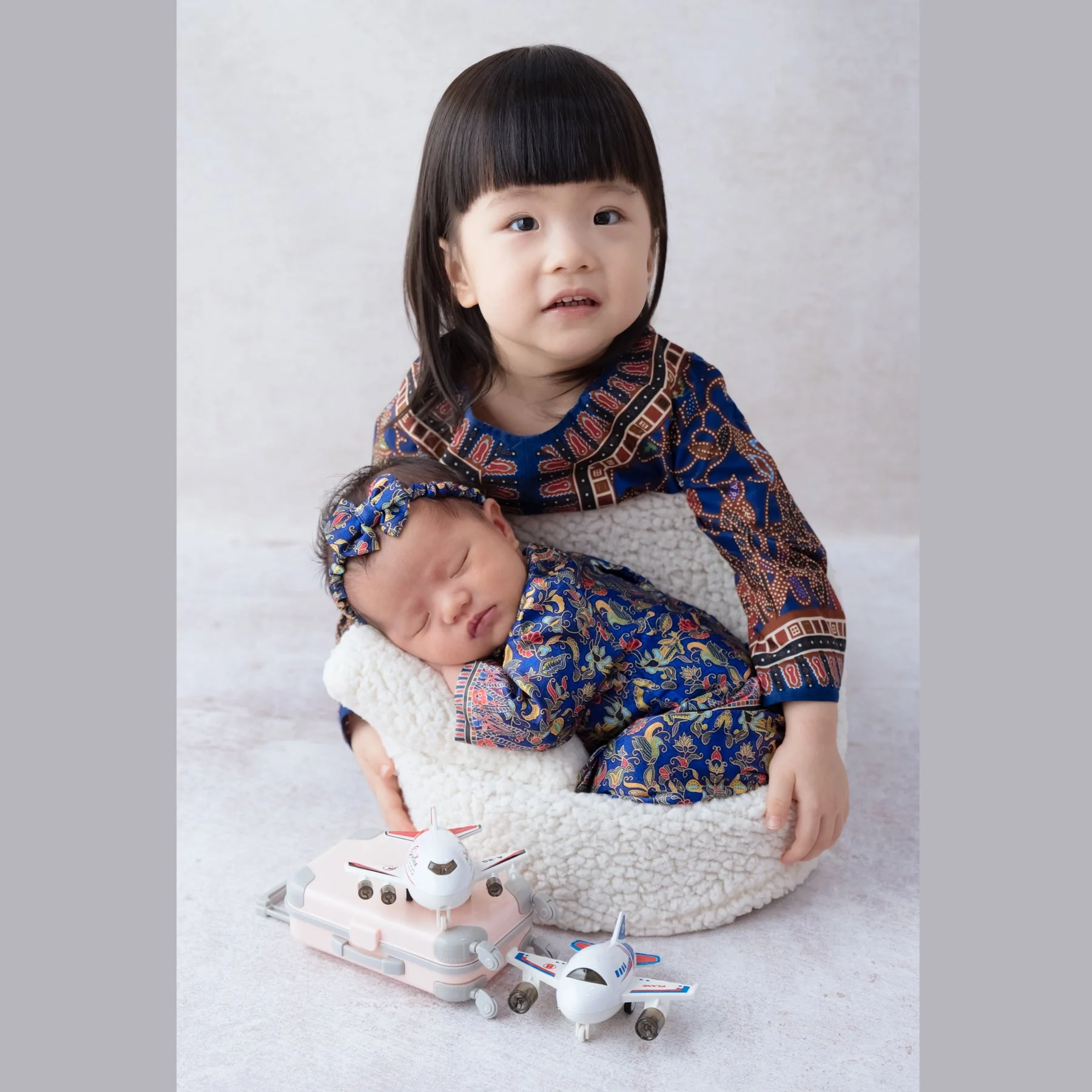 Older sibling in a patterned blue outfit sitting behind a sleeping newborn in a matching dress with toy airplanes in front