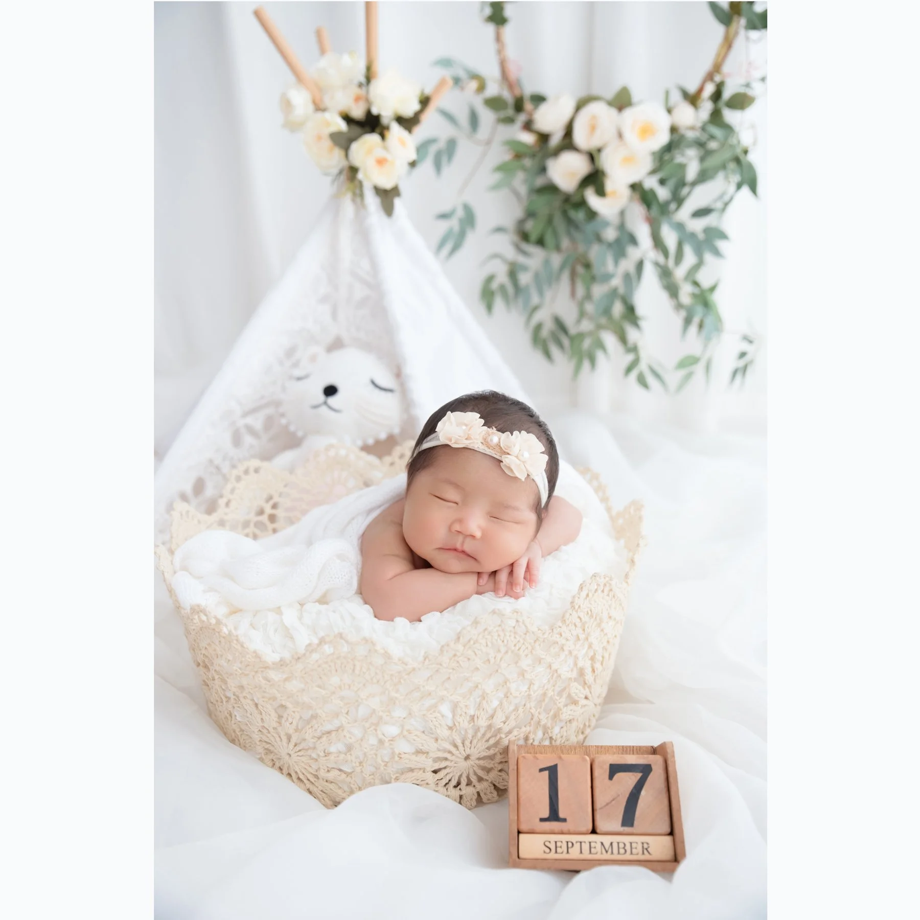 Sleeping newborn wearing a floral headband with chin on folded arms in a woven basket beneath white flowers and greenery