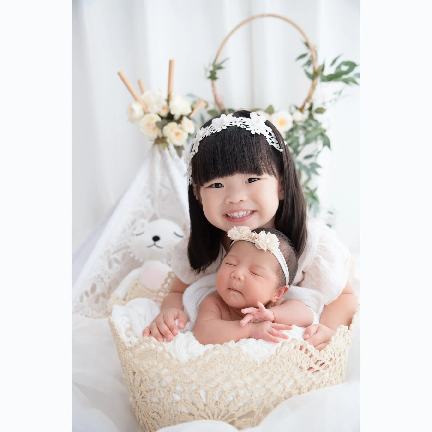 Smiling older sister embracing a sleeping newborn with a floral headband in a woven basket beneath white flowers and greenery