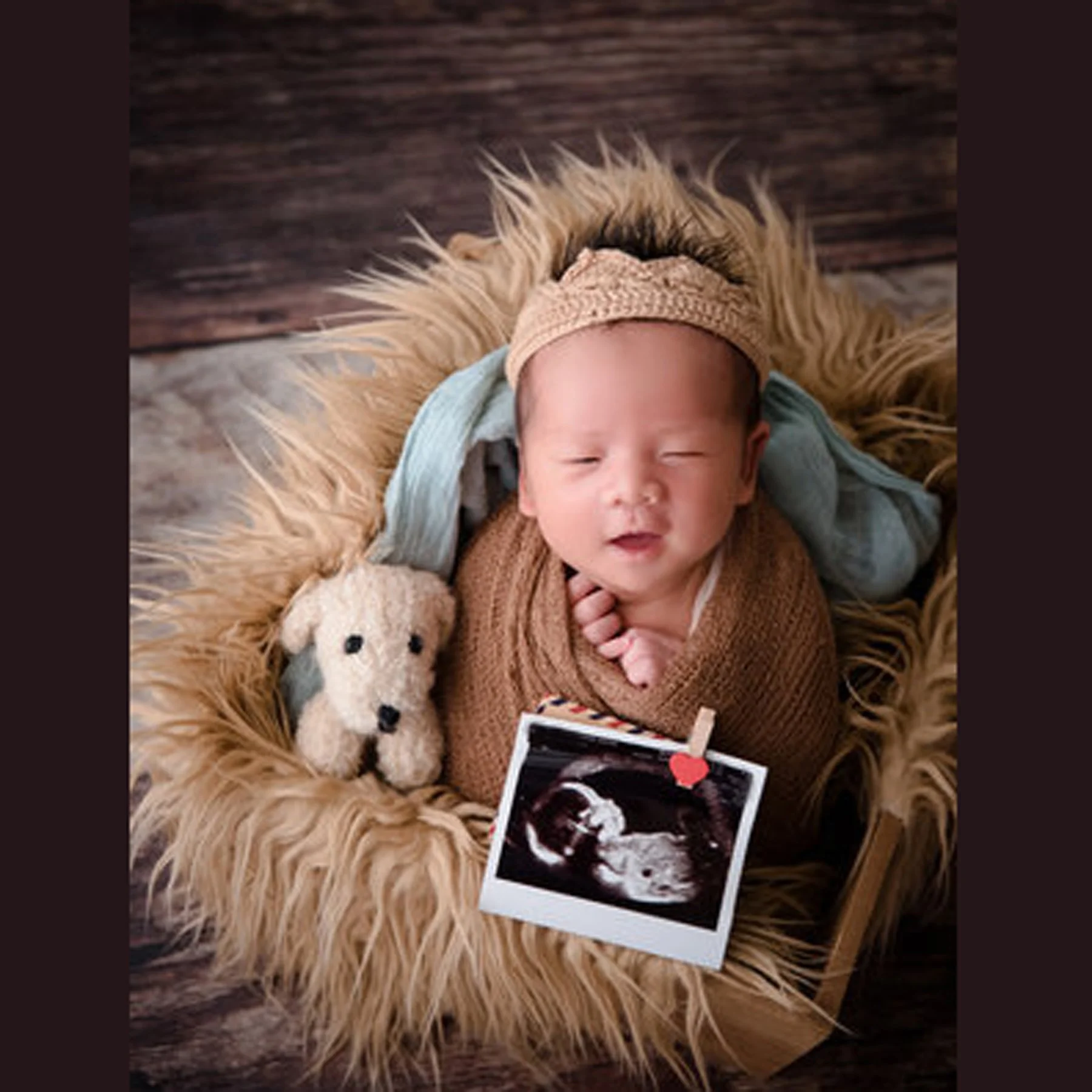 Sleeping newborn in a brown knitted wrap and beige hat nestled in a brown fur-lined basket beside a teddy bear and ultrasound picture
