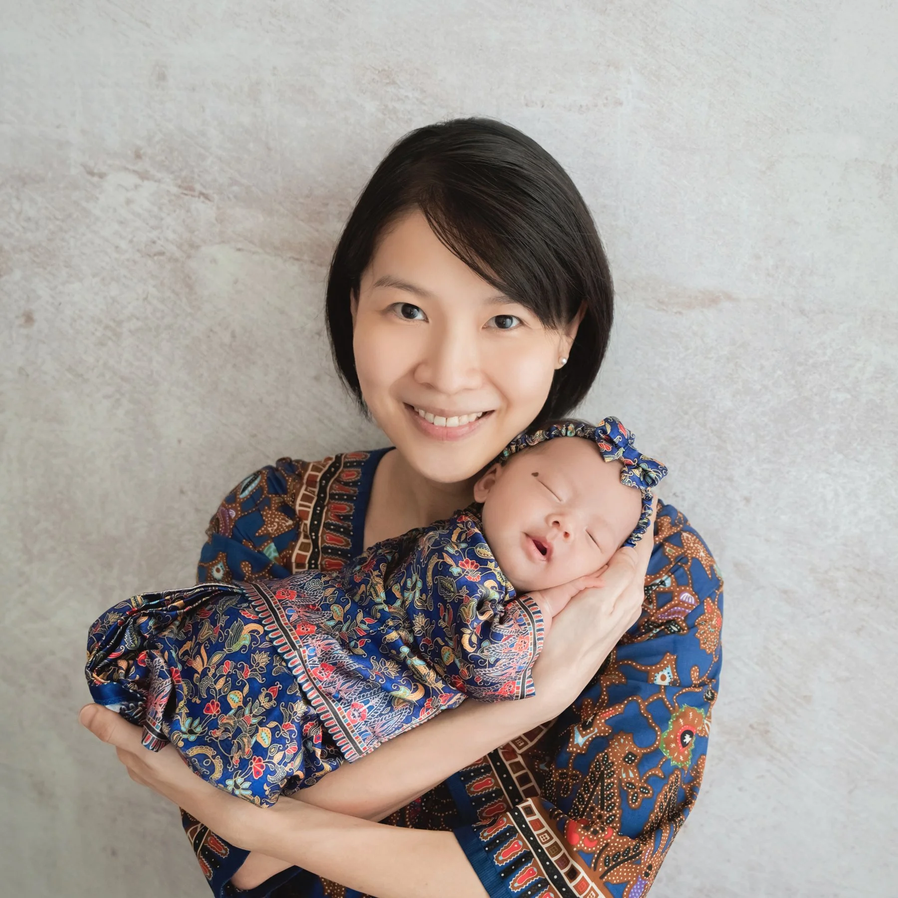 Smiling mother in a patterned blue outfit cradling a sleeping newborn in a matching dress and headband against a pale backdrop
