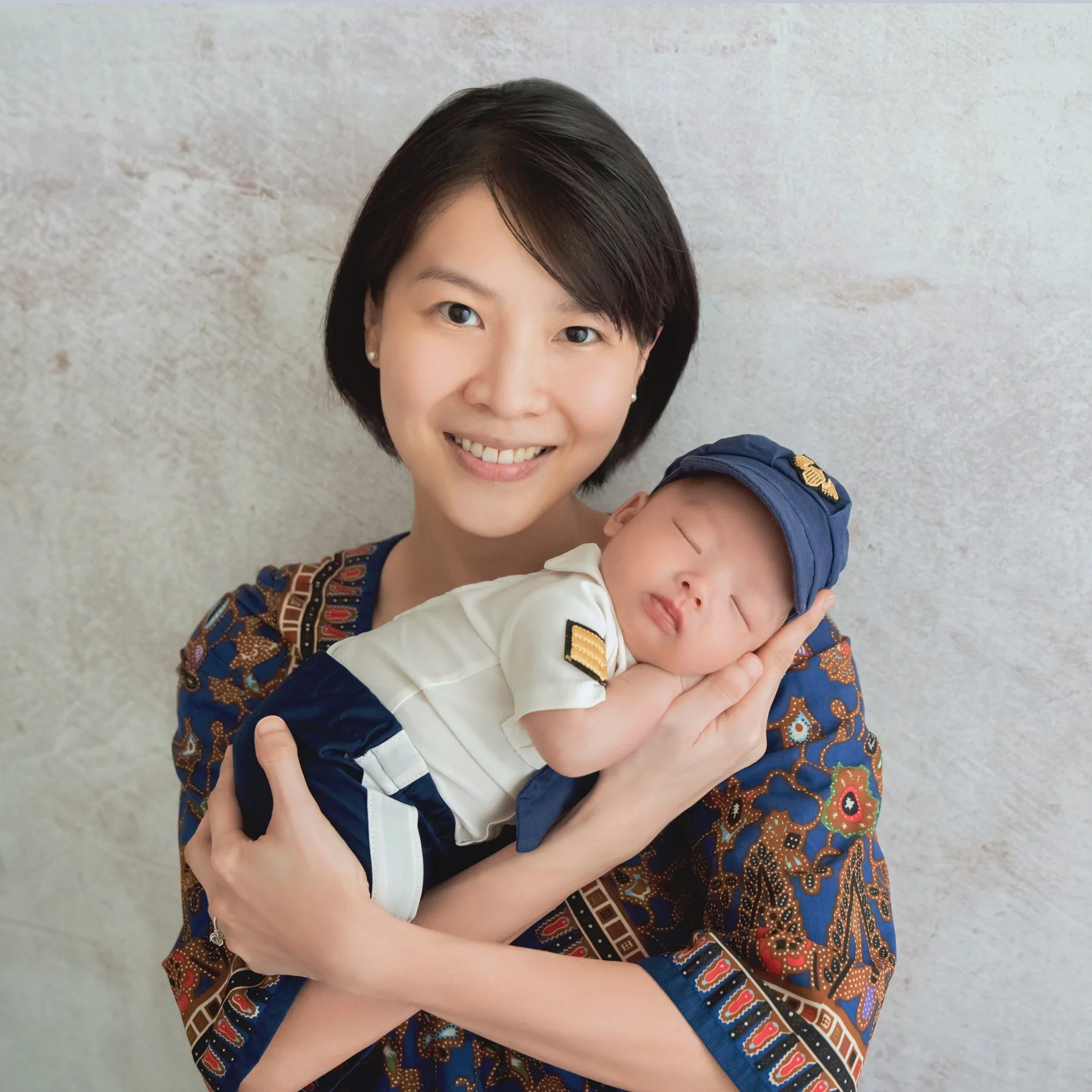 Smiling mother in a patterned blue outfit cradling a sleeping newborn in a white pilot shirt and navy cap