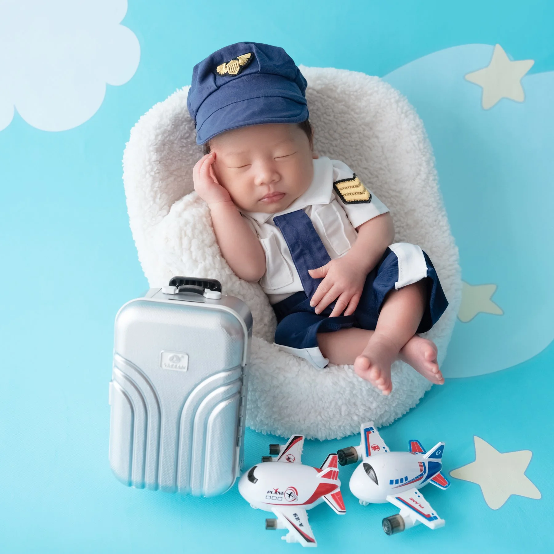 Sleeping newborn in a navy pilot cap and white uniform reclining in a basket beside a silver suitcase and toy airplanes