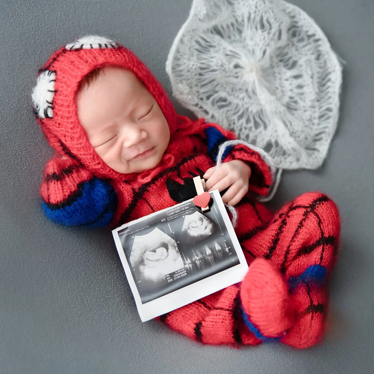 Sleeping newborn in a knitted Spider-Man costume lying on one side beside a white web prop