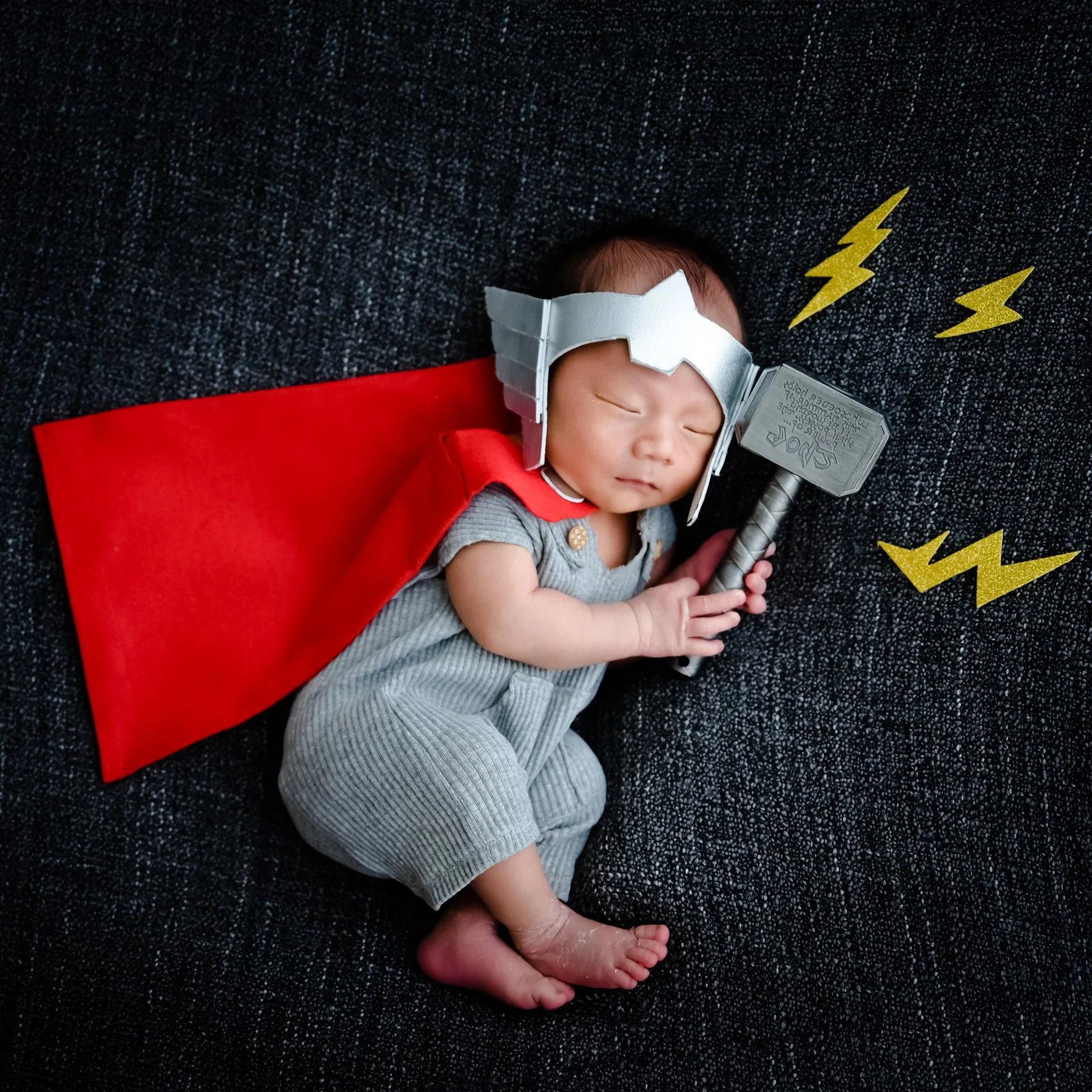 Sleeping newborn dressed as Thor, curled on one side holding a hammer with a red cape and lightning props