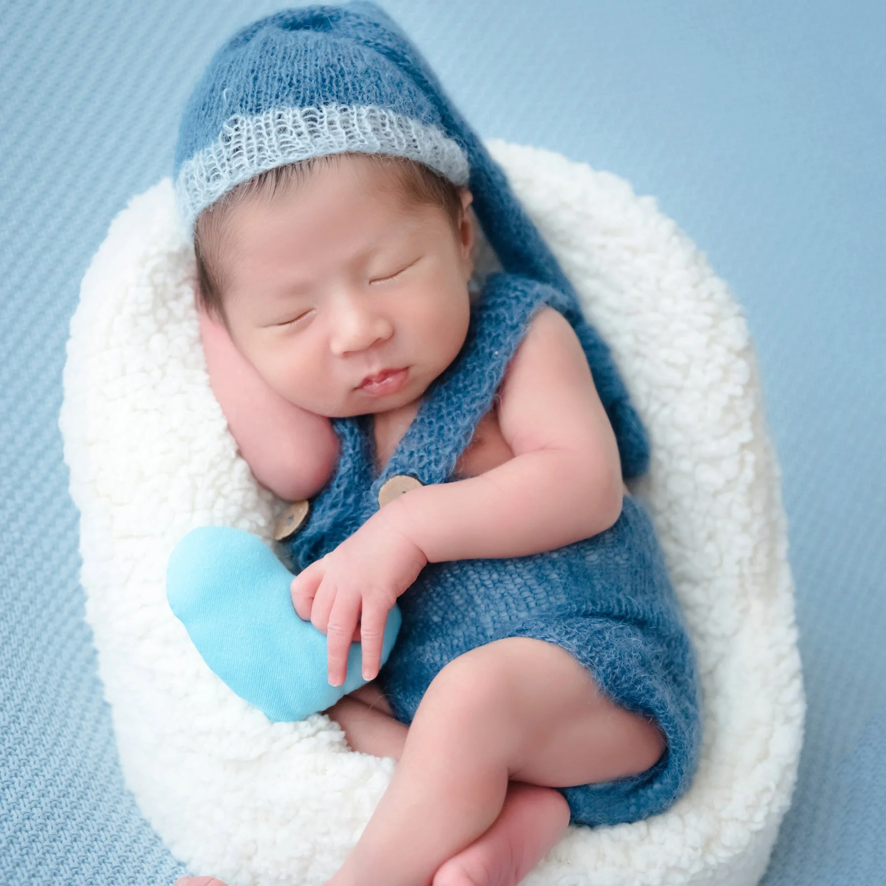 Sleeping newborn in blue knitted overalls and a matching hat curled in a white basket with a blue heart-shaped cushion