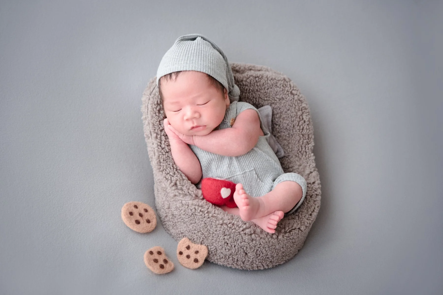 Sleeping newborn in a grey knitted hat and romper curled on one side in a soft grey basket with a red toy cup and miniature cookies nearby