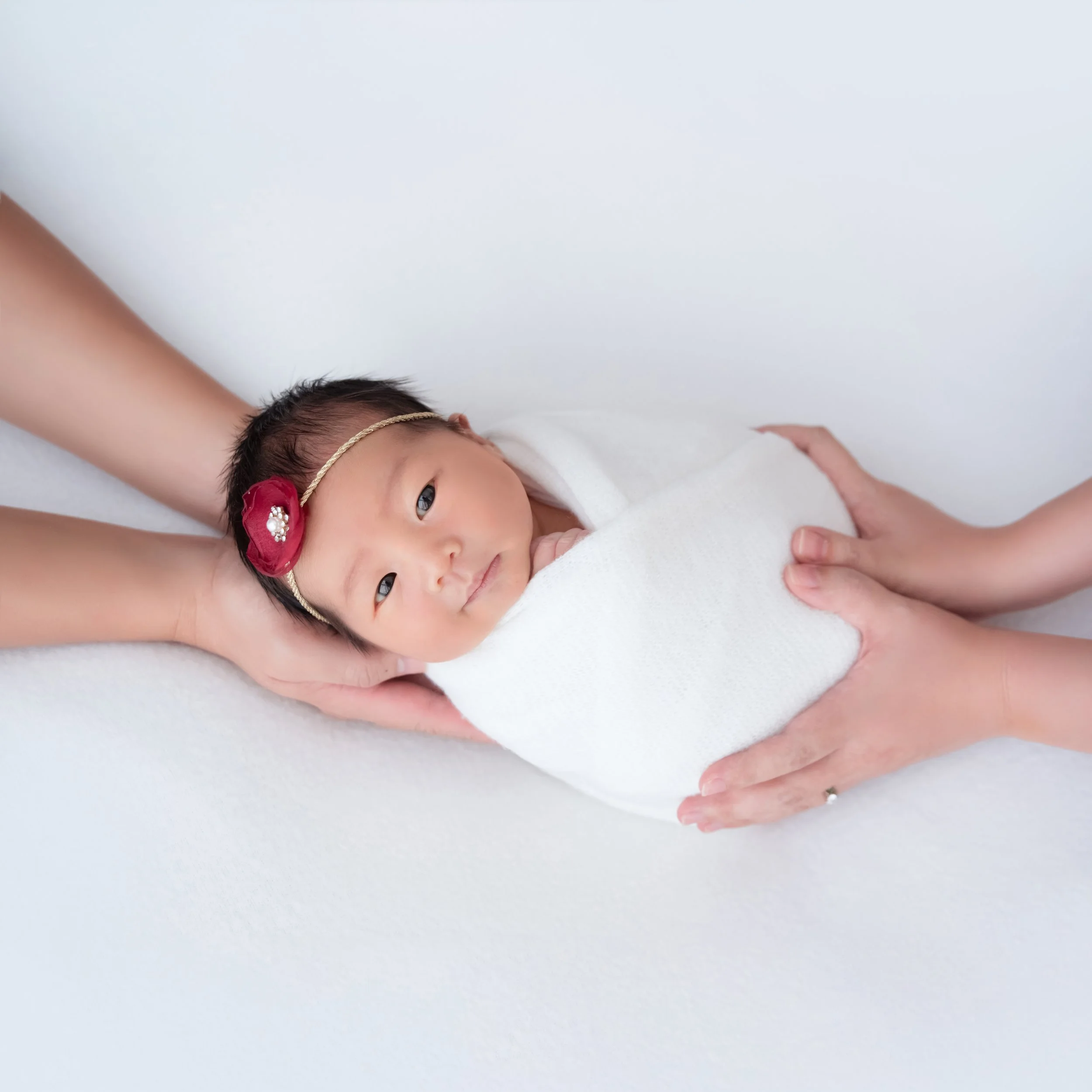 Sleeping newborn in a white swaddle and colourful flower headband supported by adult hands against a pale backdrop
