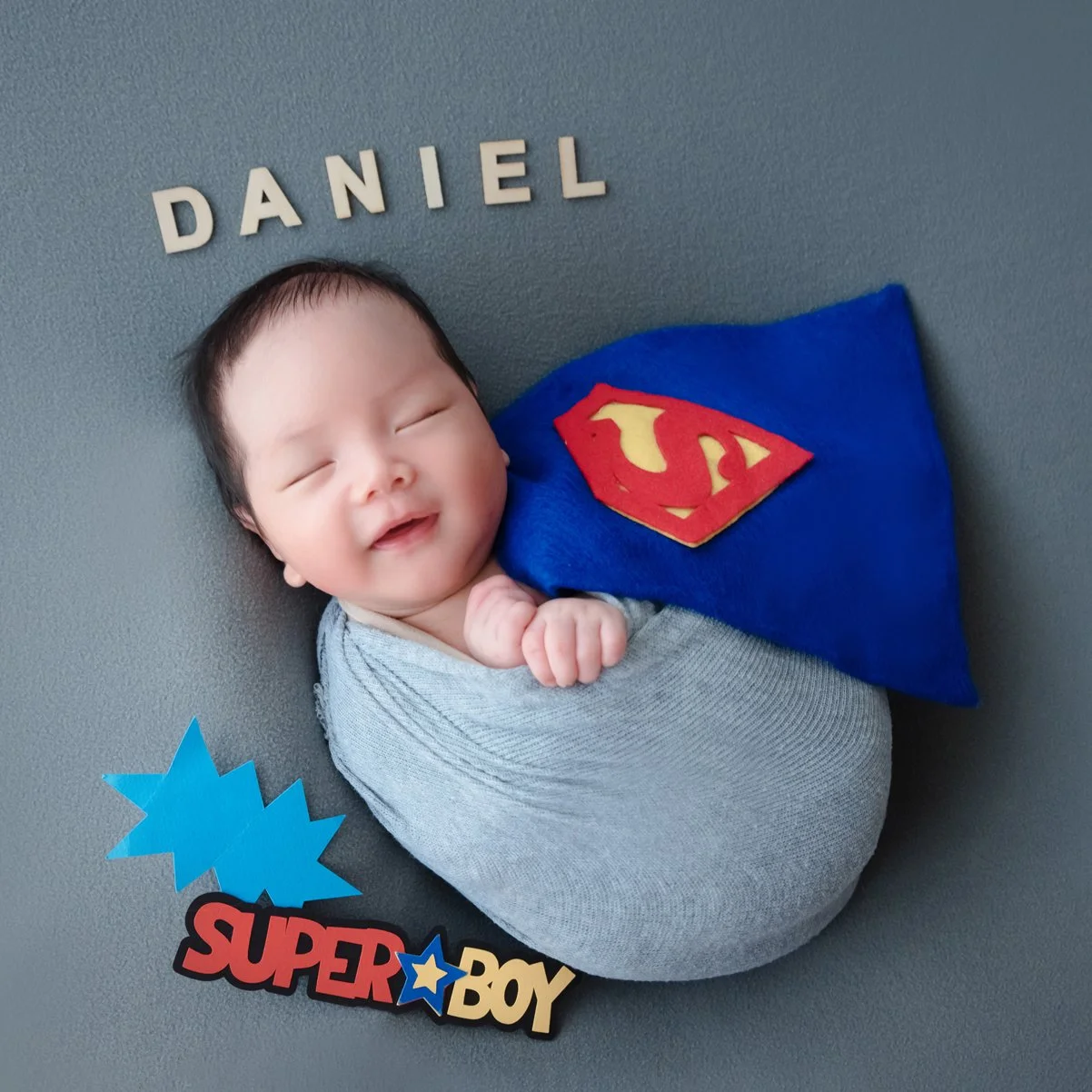 Newborn wrapped in grey fabric beside a blue Superman cushion and colourful comic-style props