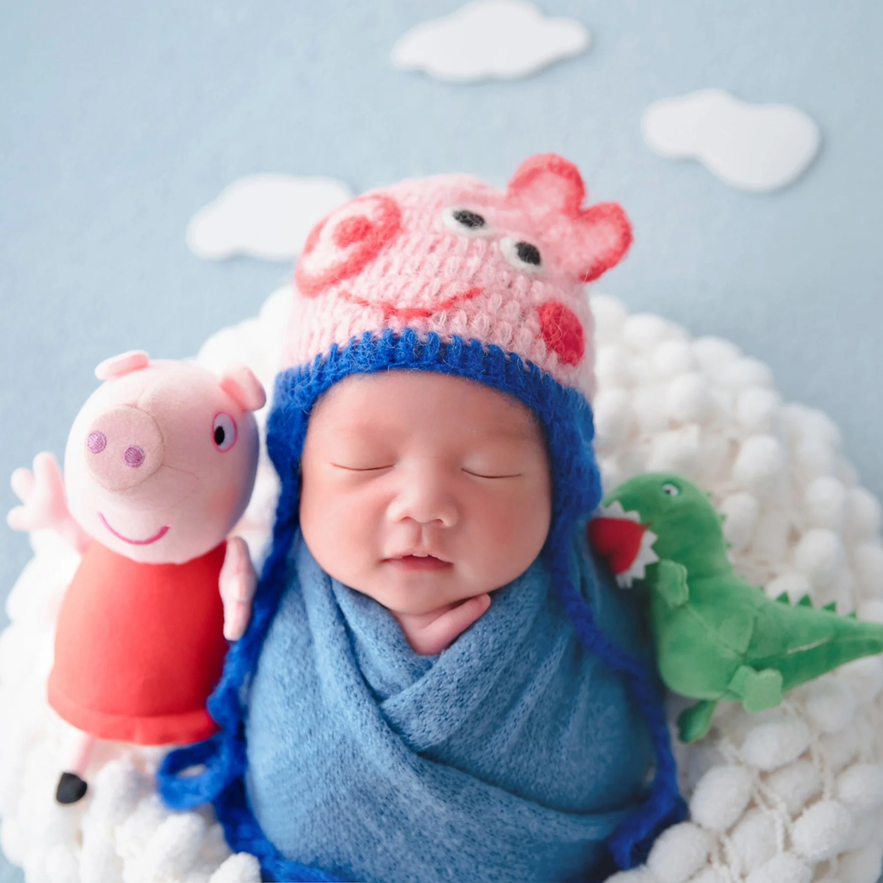 Newborn in a blue swaddle and George Pig hat resting in a fluffy white basket beside pig and dinosaur toys