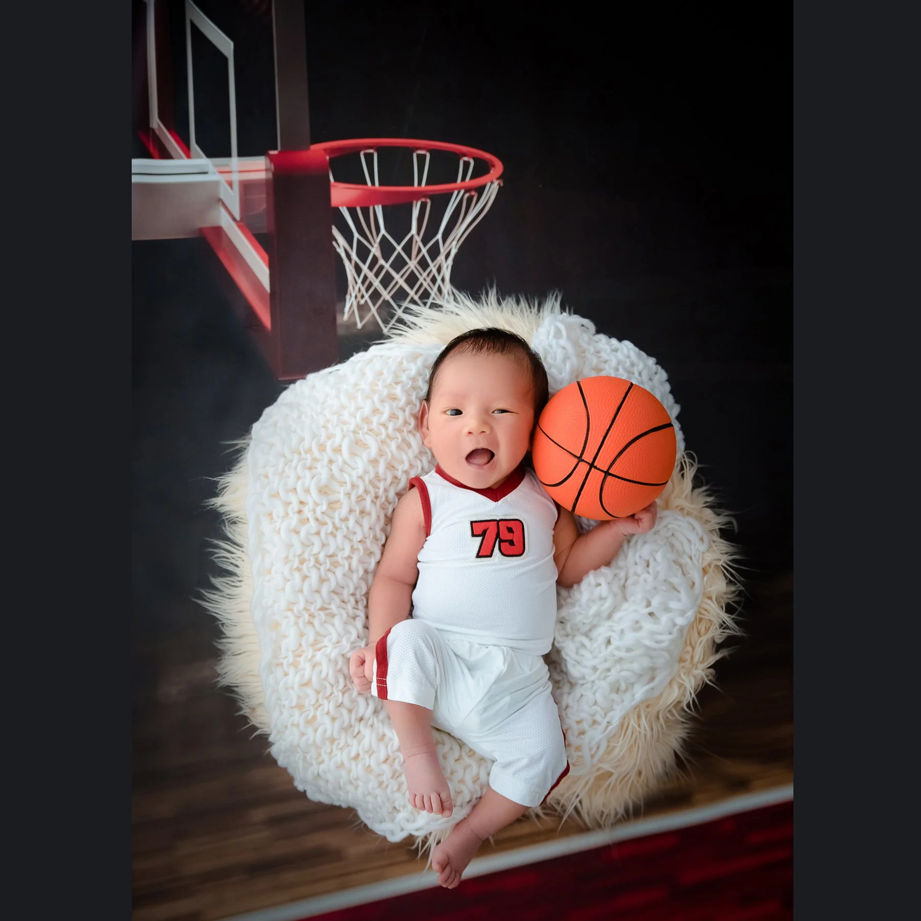 Yawning newborn in a white basketball outfit reclining in a fluffy white basket beside an orange basketball and toy hoop