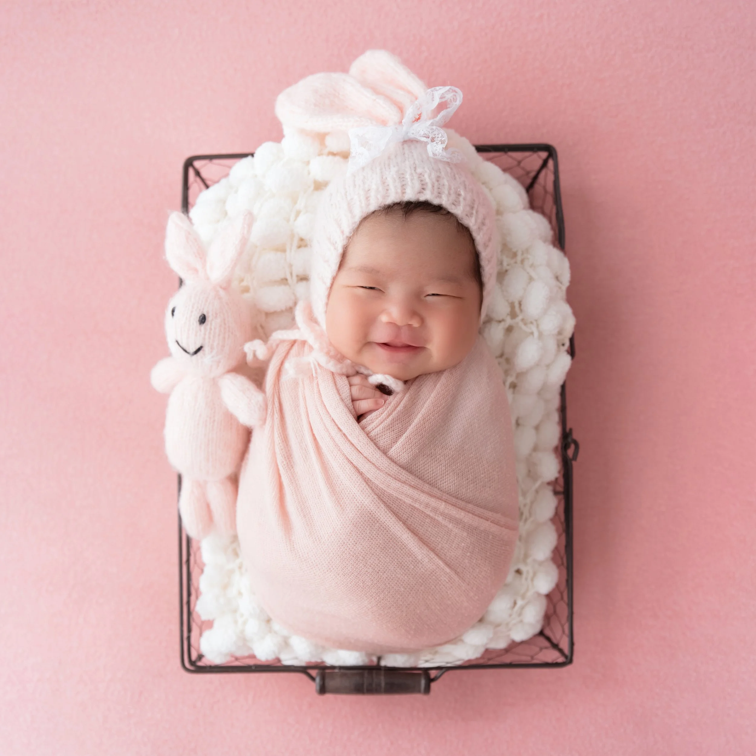 Sleeping newborn in a pale pink wrap and knitted bunny hat nestled in a white-lined wire basket against pink