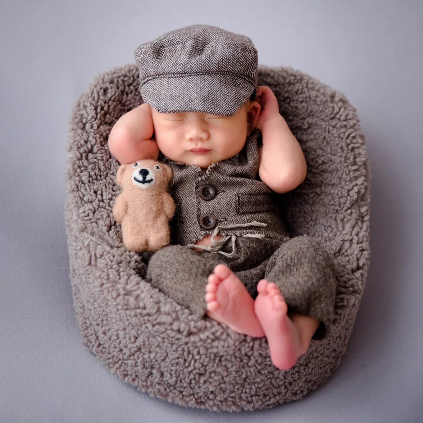 Sleeping newborn in a grey flat cap and brown knitted outfit reclining in a soft grey basket with a teddy bear and one hand beside the head