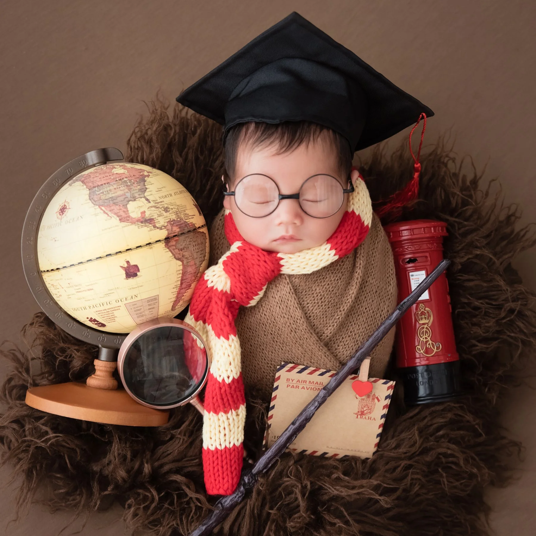 Sleeping newborn in round glasses, a graduation cap and red-and-cream scarf beside a globe and toy wand